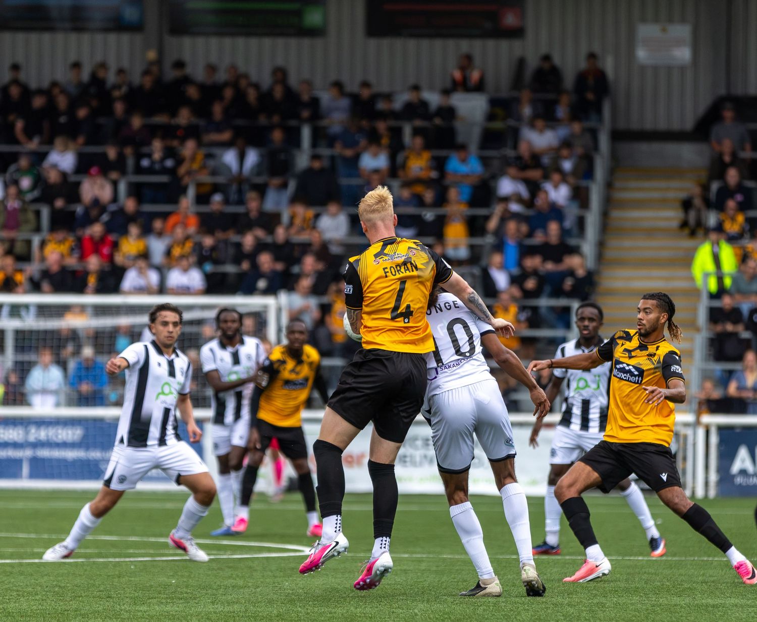Football players compete for a header during a match, with fans watching from the stands in the background.