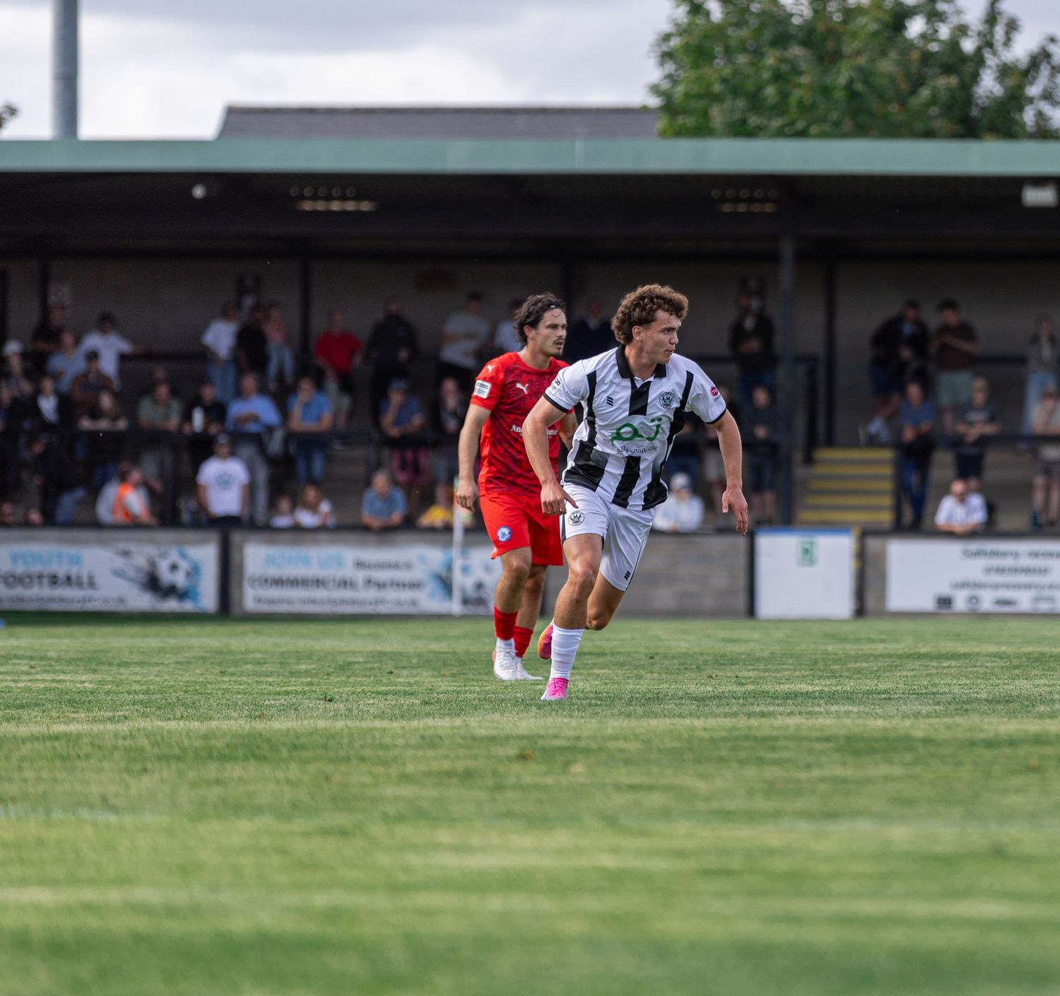 Two football players in black-white and red kits compete on a green pitch with spectators watching from stands.