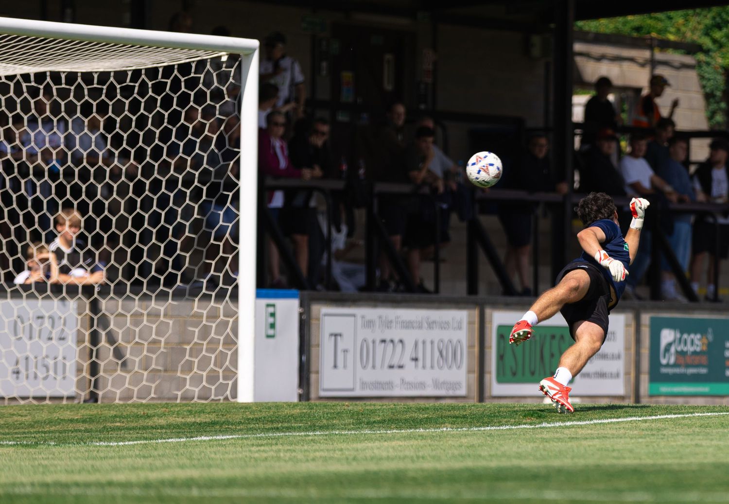 Soccer player kicks ball toward goal net during outdoor match with spectators watching from stands.