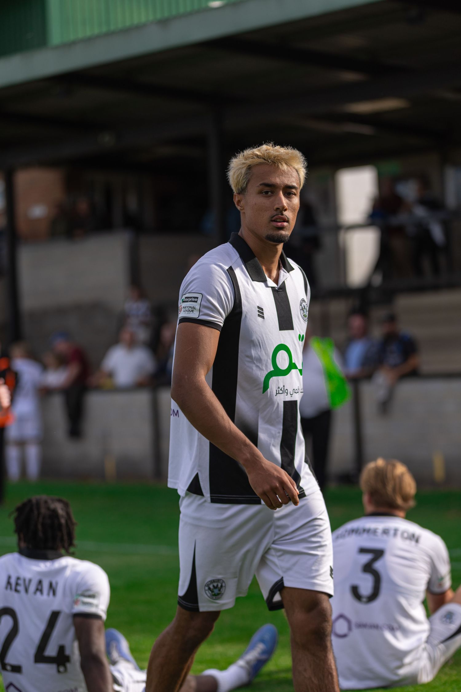 Blonde soccer player in black and white kit stands on field during match, with teammates wearing numbers 24 and 3 nearby.