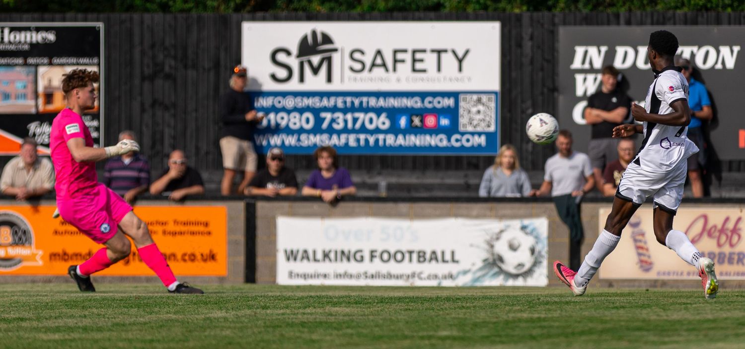Two soccer players compete for the ball during an outdoor match near SM Safety Training advertising boards.