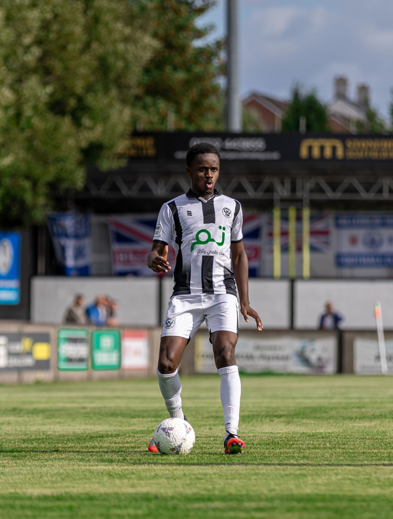 Soccer player in black and white kit dribbling ball on green pitch during sunny match day.