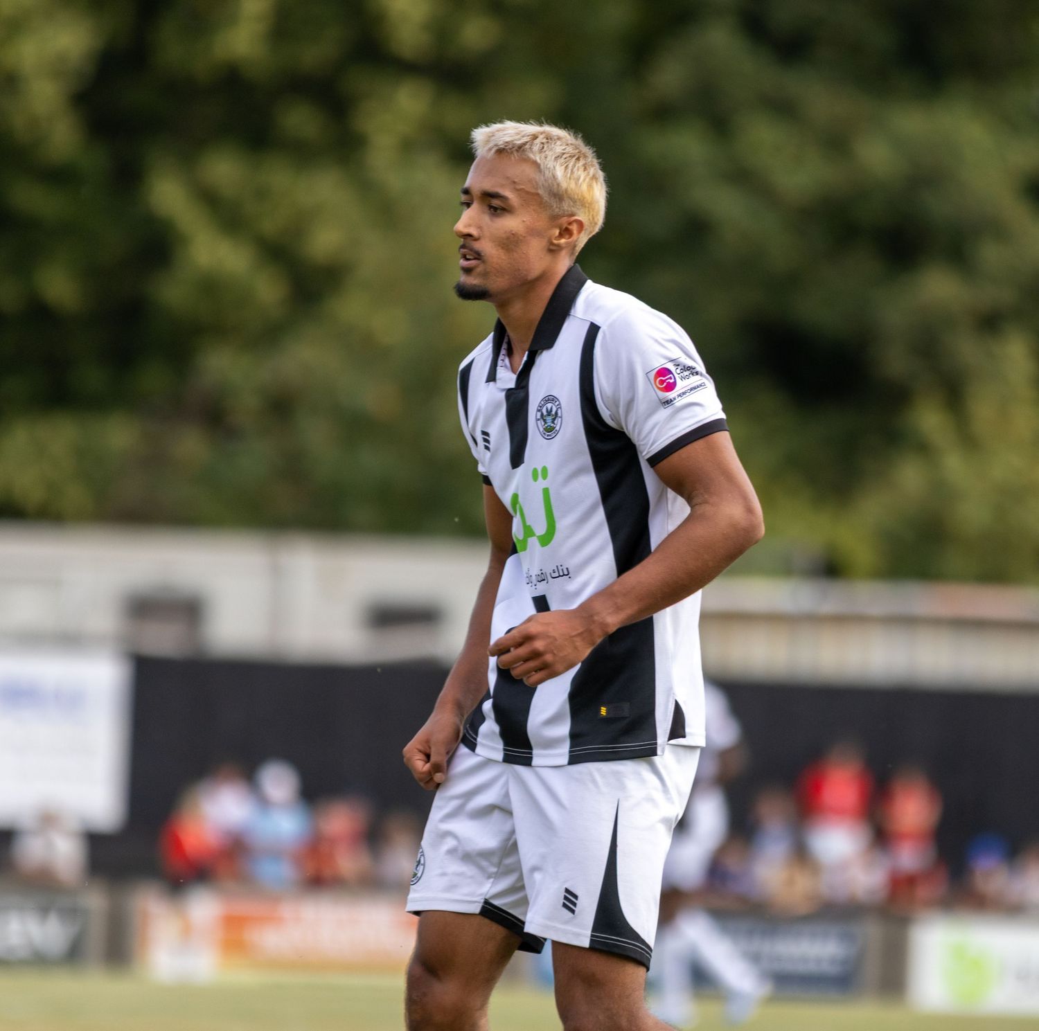 Close-up of a footballer in black and white striped kit with blonde highlights standing on a pitch.