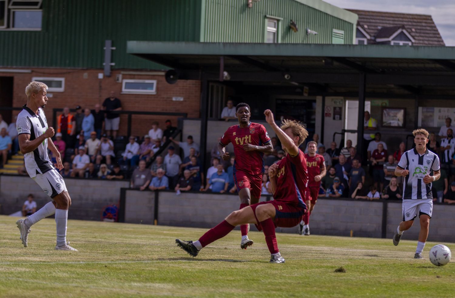 A red-kitted player shoots at goal during a non-league football match with defenders nearby.