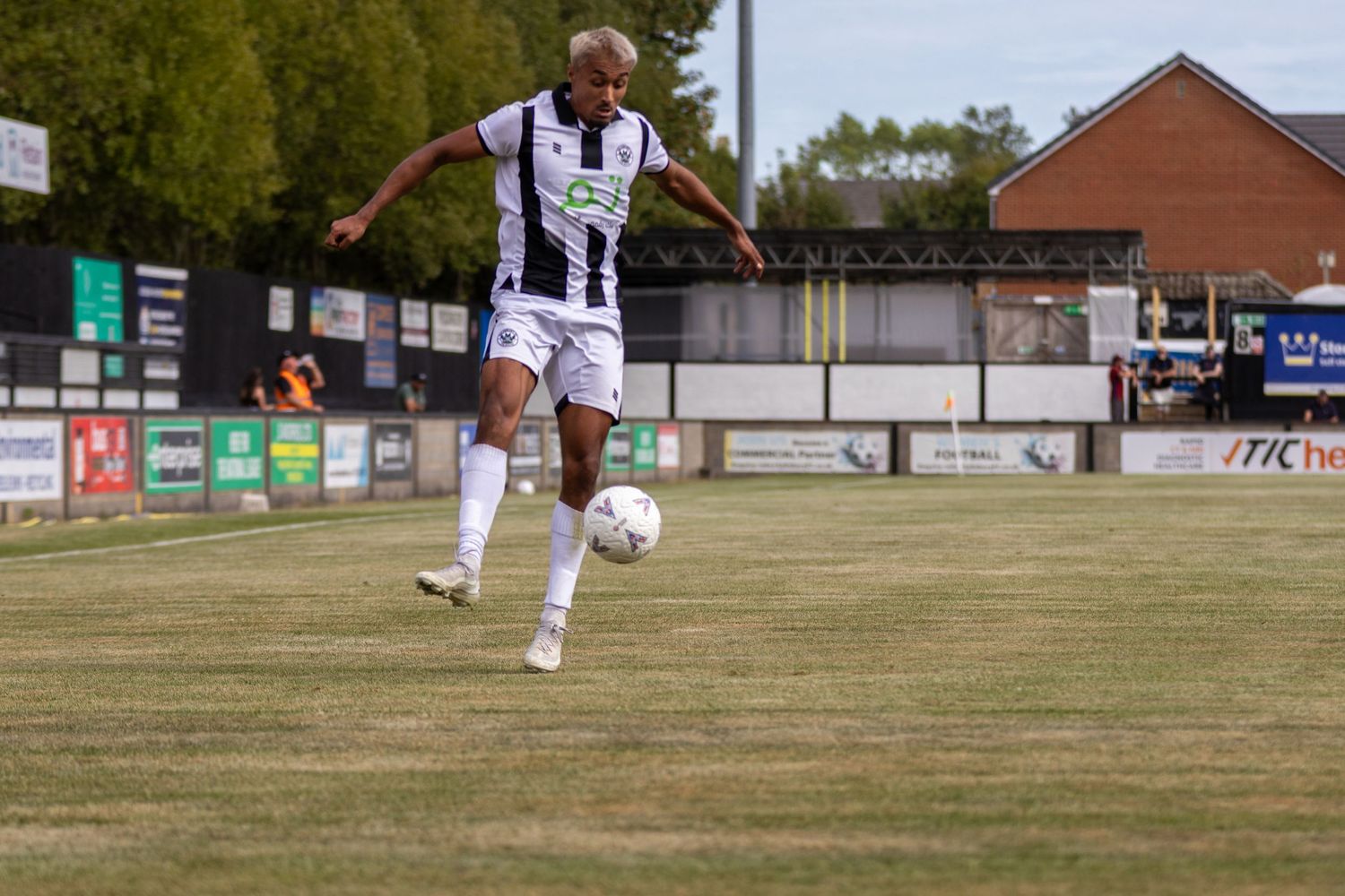 Soccer player in black and white kit strikes ball on green pitch with advertising boards in background.