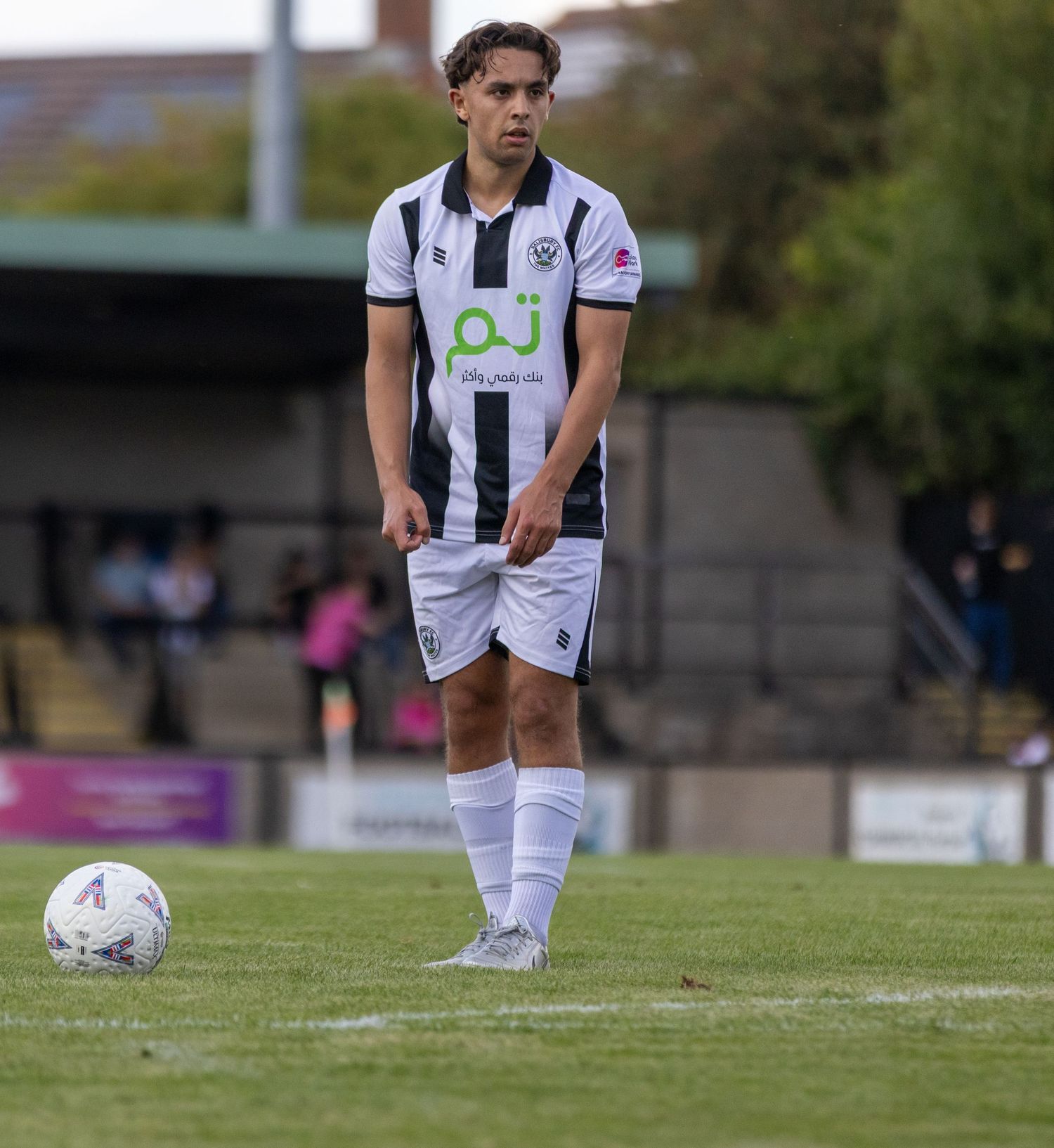 A footballer in a black and white striped jersey stands focused near the ball on a pitch.