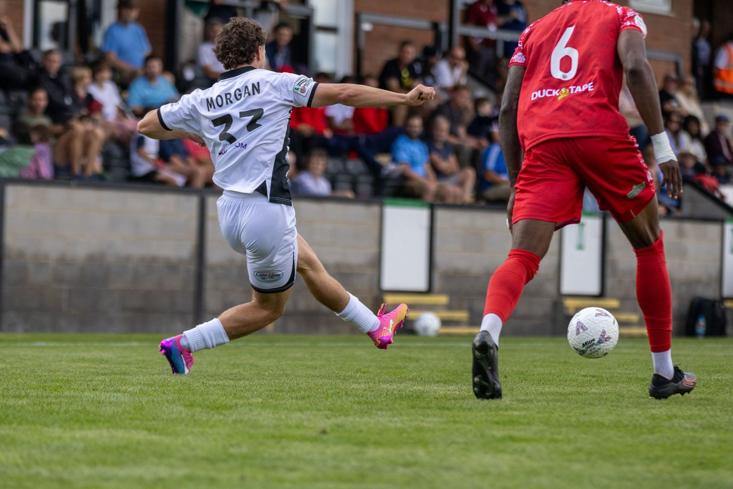Footballer Holigan in white kit shoots the ball while a red-kitted number 6 opponent looks on.