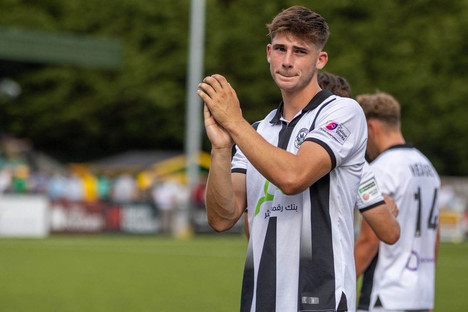 A young male footballer in black and white striped kit claps fans after a sunny day match.