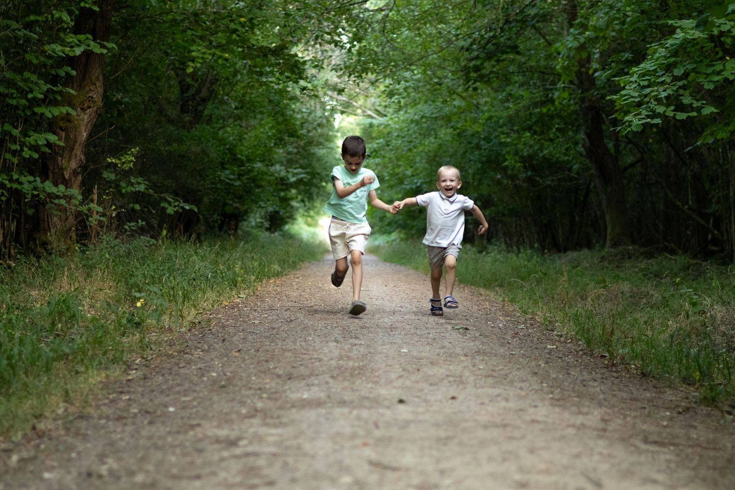 Two kids hold hands and run joyfully along a dirt path through a green forest.