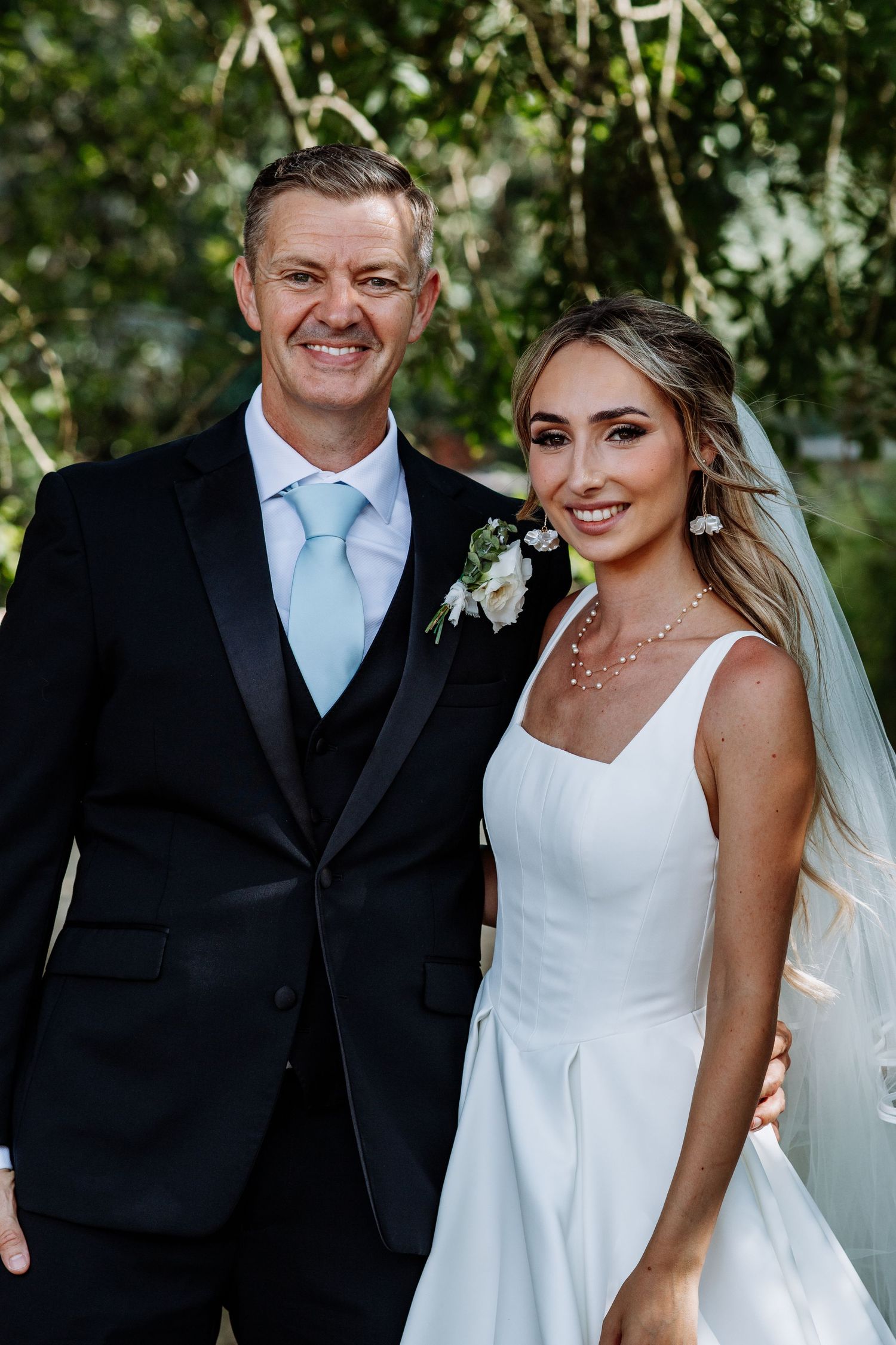 Couple at outdoor wedding, man in black tuxedo with blue tie and bride in elegant white gown smiling.