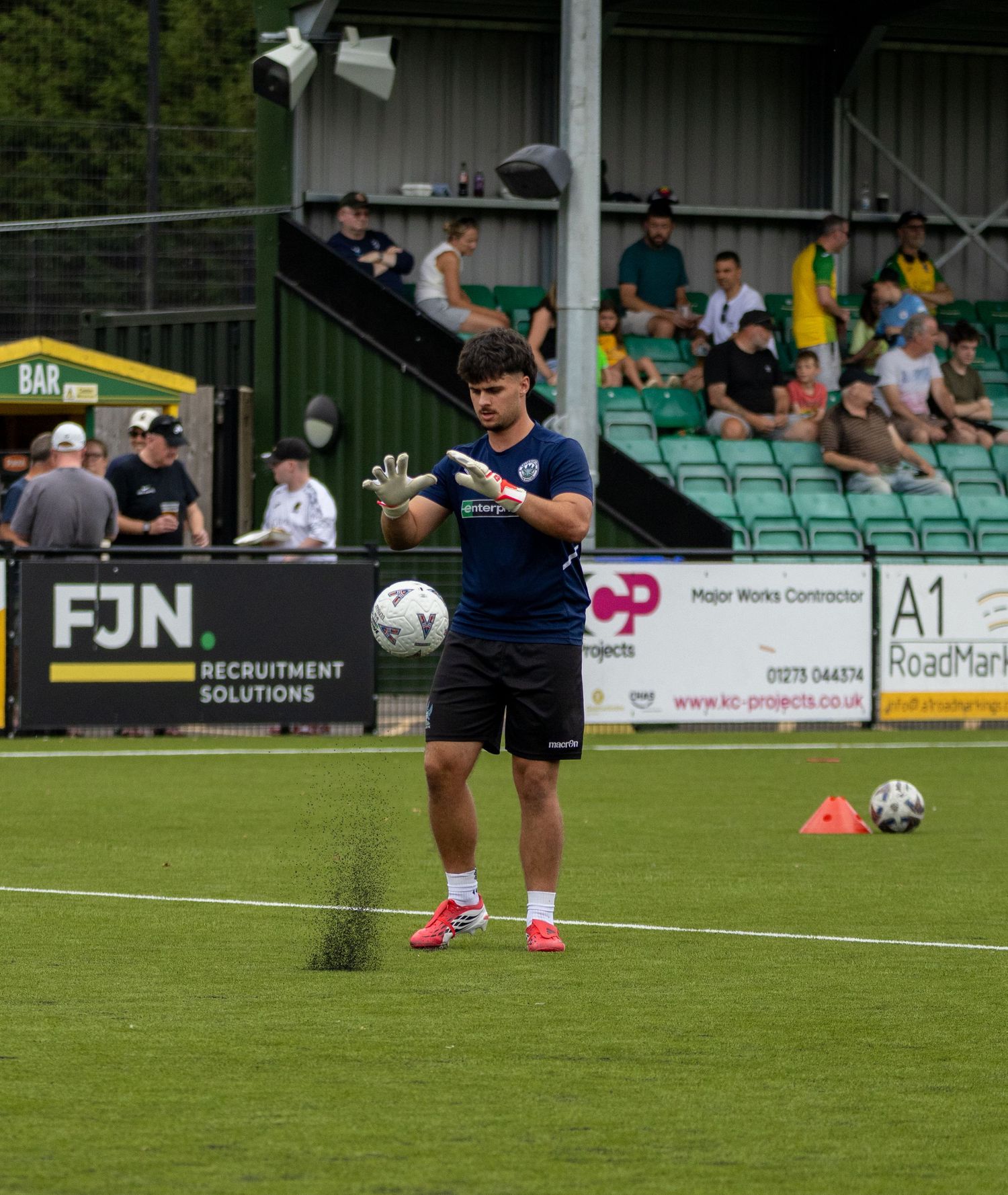 Football goalkeeper coach in navy kit tossing ball during training warmup at stadium with FJN sponsor board visible.