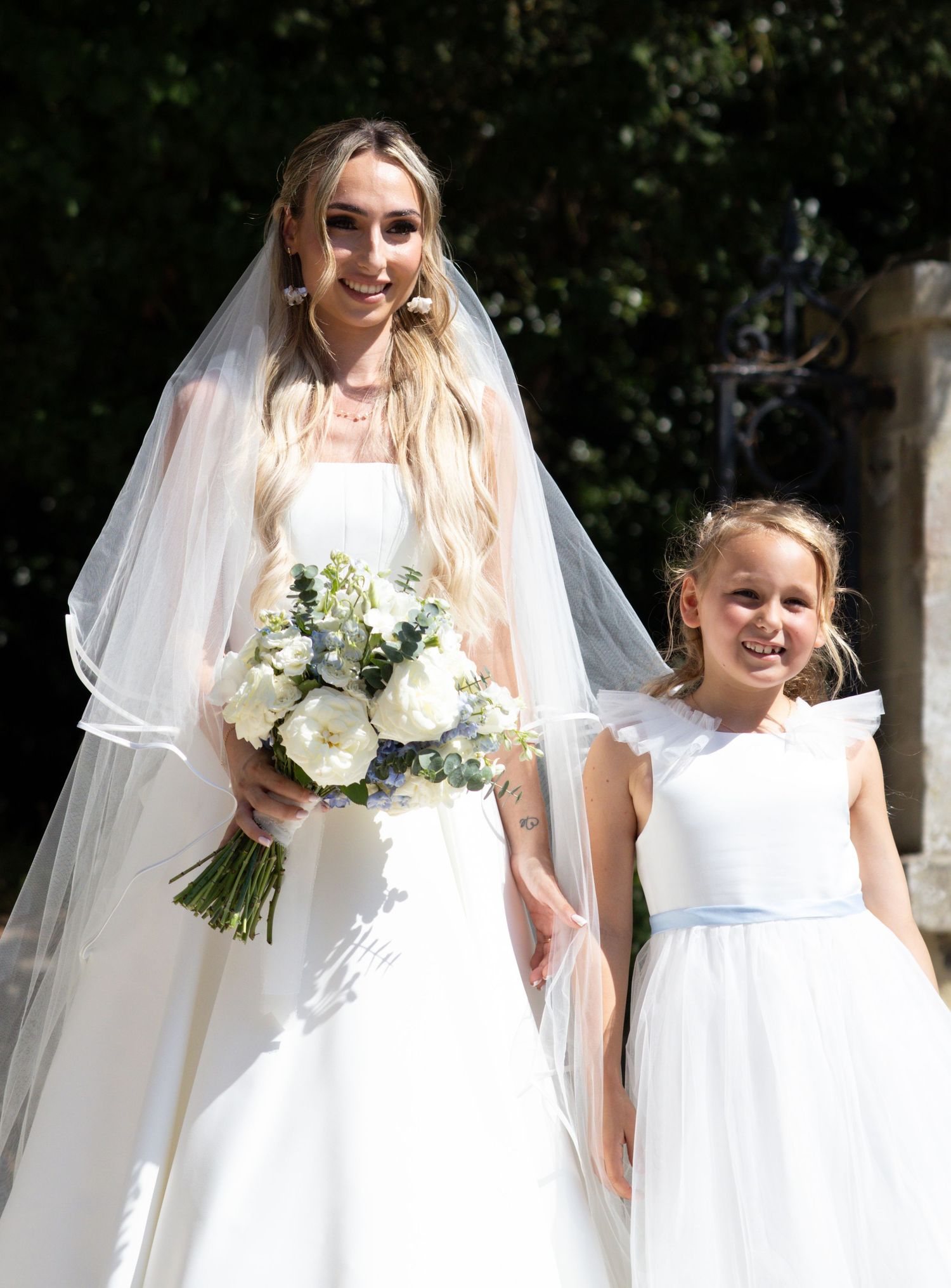Bride in white gown with veil holding bouquet stands with young flower girl in white dress outdoors.