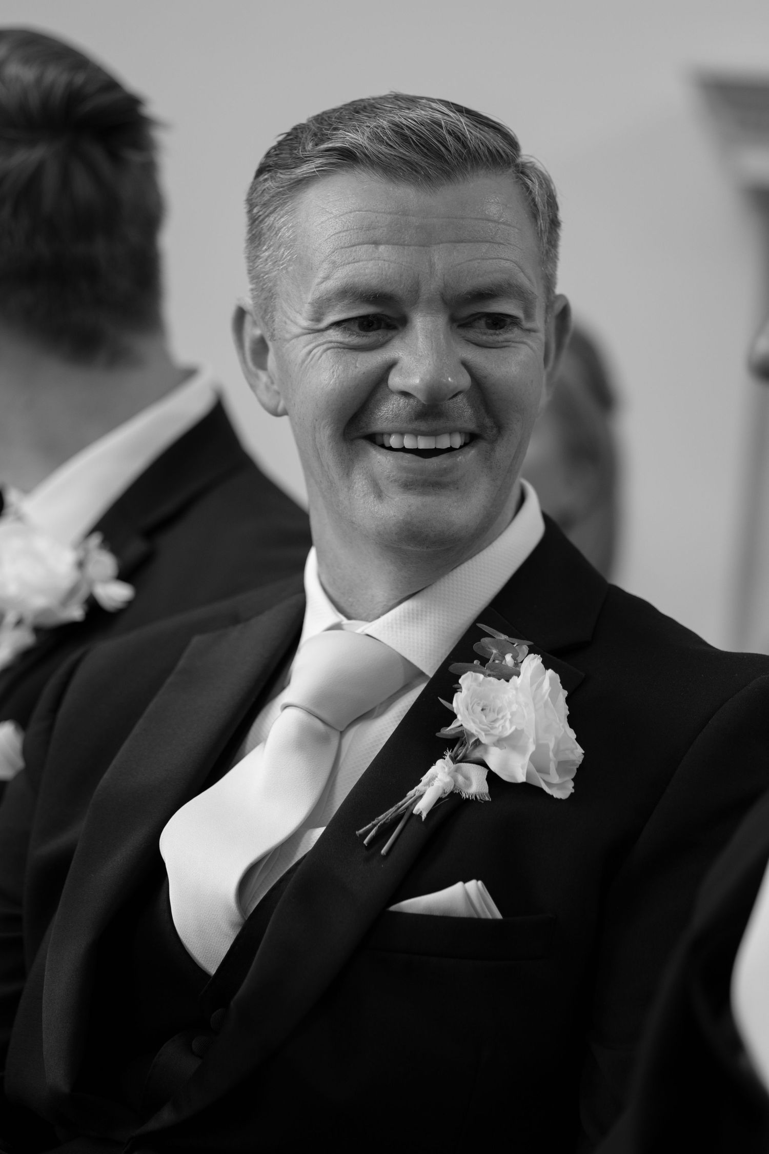 Smiling groom in black tuxedo with white rose boutonniere and pink tie at wedding celebration.