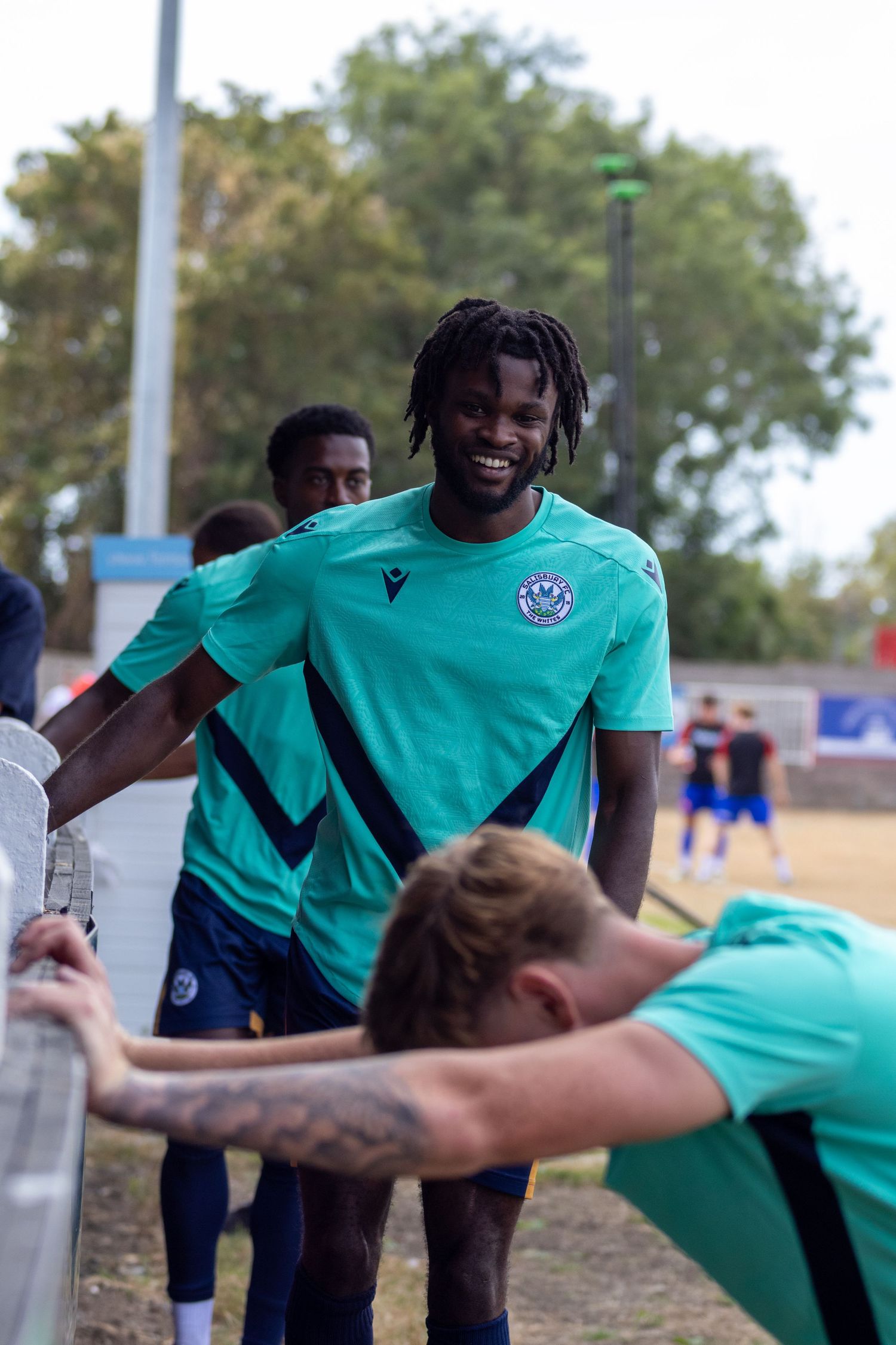 Smiling football player in teal kit stretching by barrier with teammates before match.