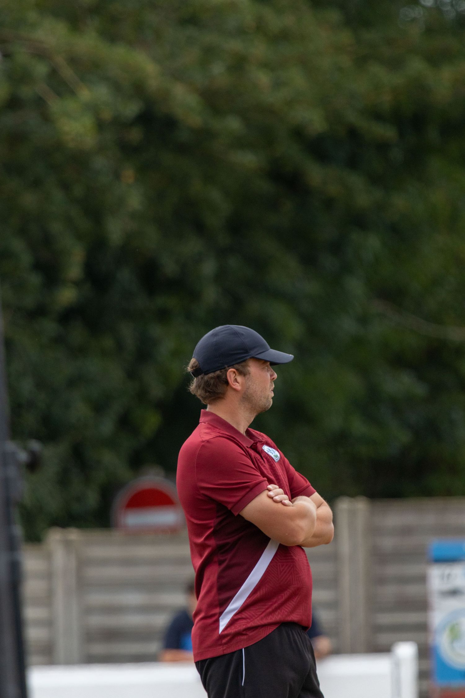 Football coach in maroon polo and cap watching match with arms crossed on touchline.
