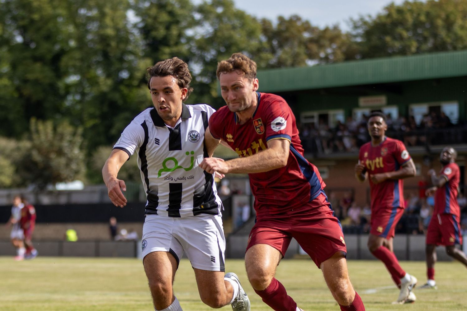 A player in black-white kit races against a red-kitted opponent during a non-league football match.