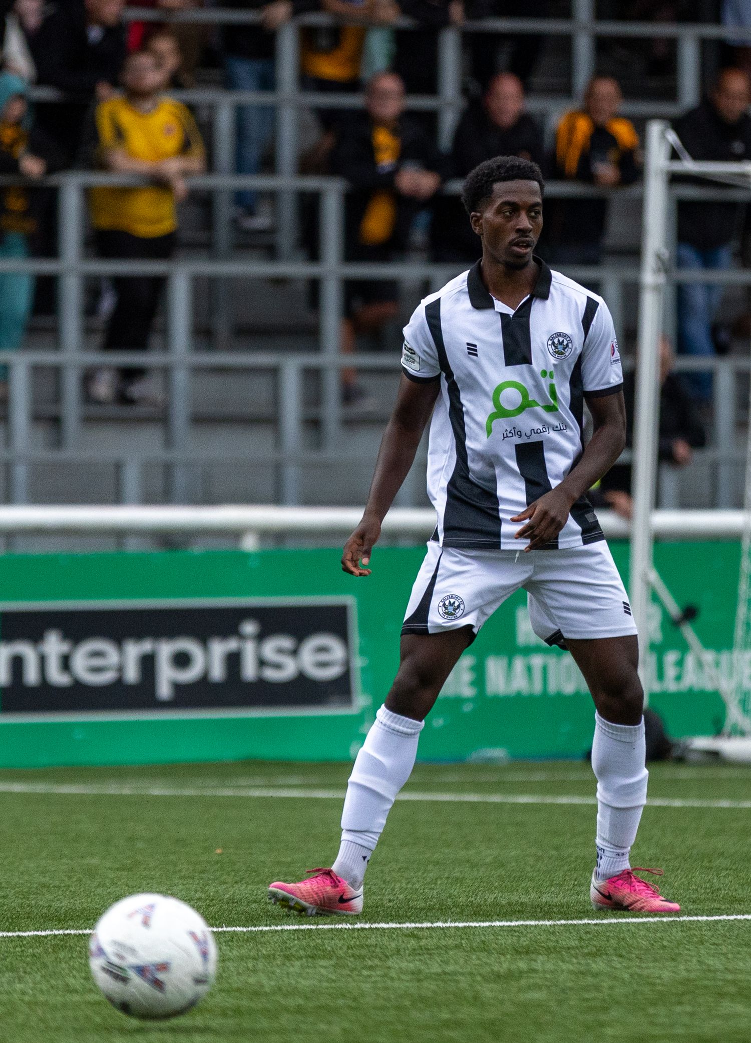 Soccer player in black and white kit with pink boots controls ball on green pitch during National League match.