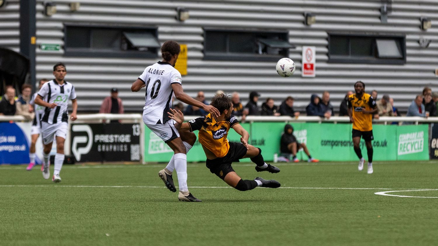 Two soccer players compete for the ball during a match, one sliding in a tackle on a green pitch.