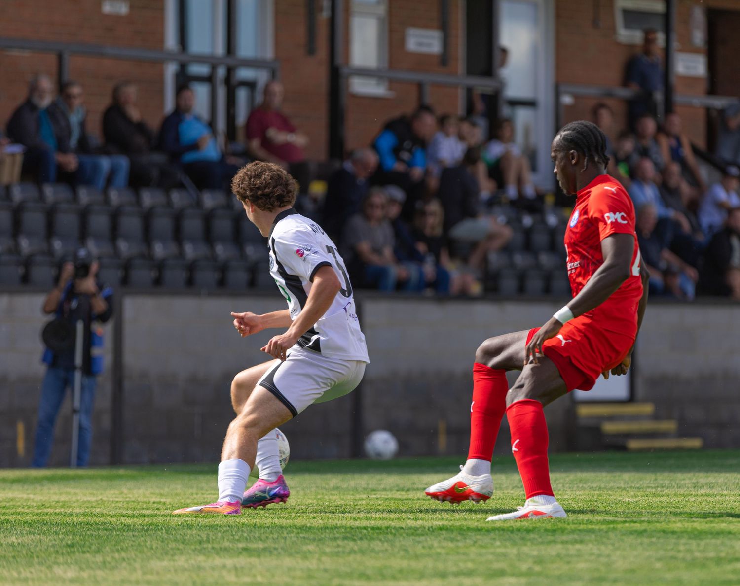 Two soccer players compete for the ball during a sunny outdoor match, one in red and one in black and white kit.