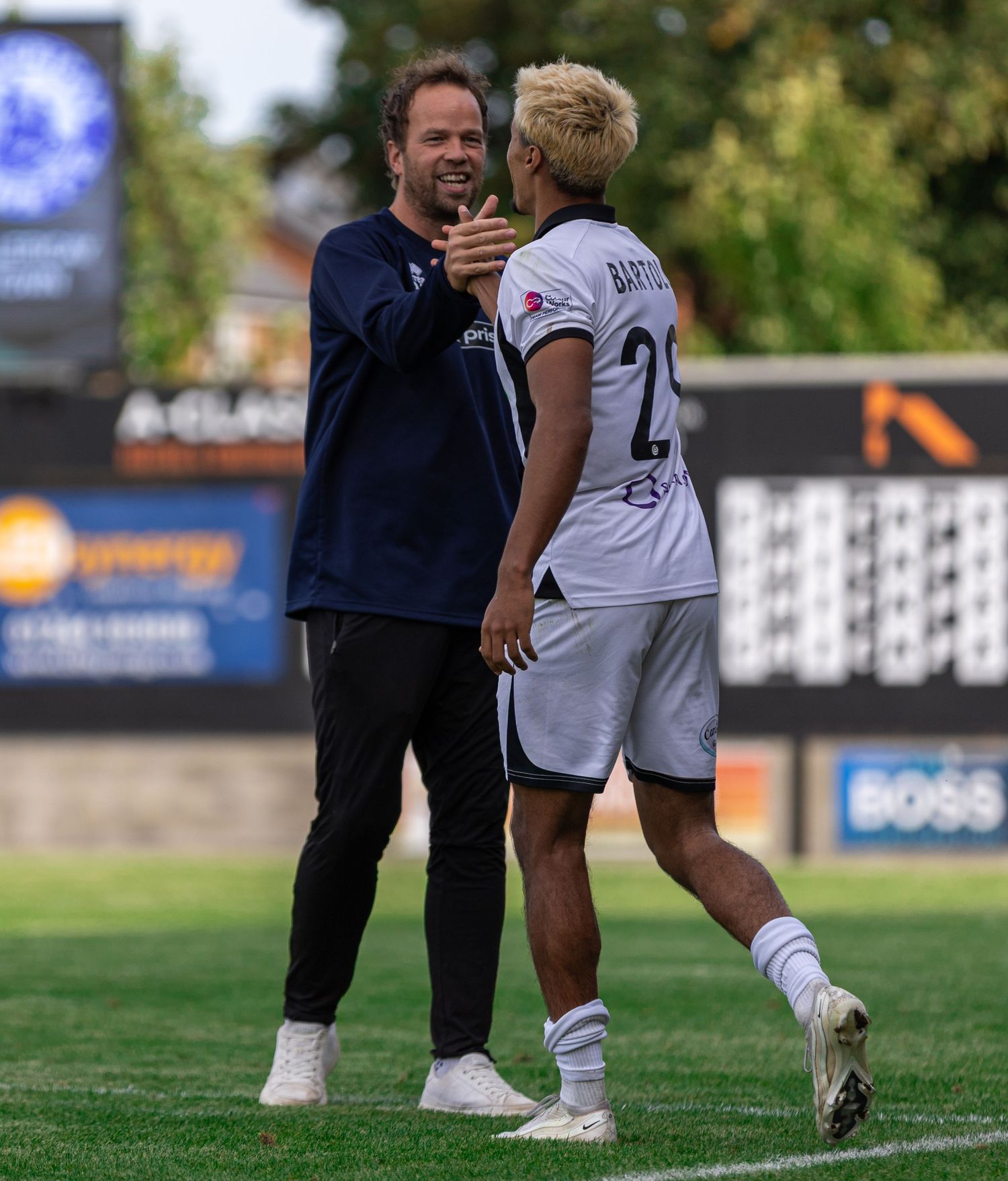 Two soccer players share a warm handshake on the field during a match, one in a dark jacket and one in a white jersey.