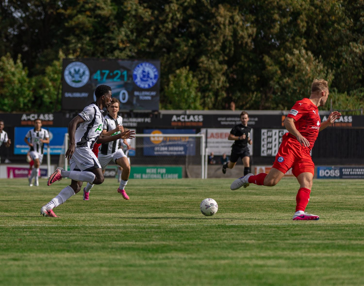 Soccer players compete on a green field during a sunny match, with a scoreboard showing 47-21 in the background.