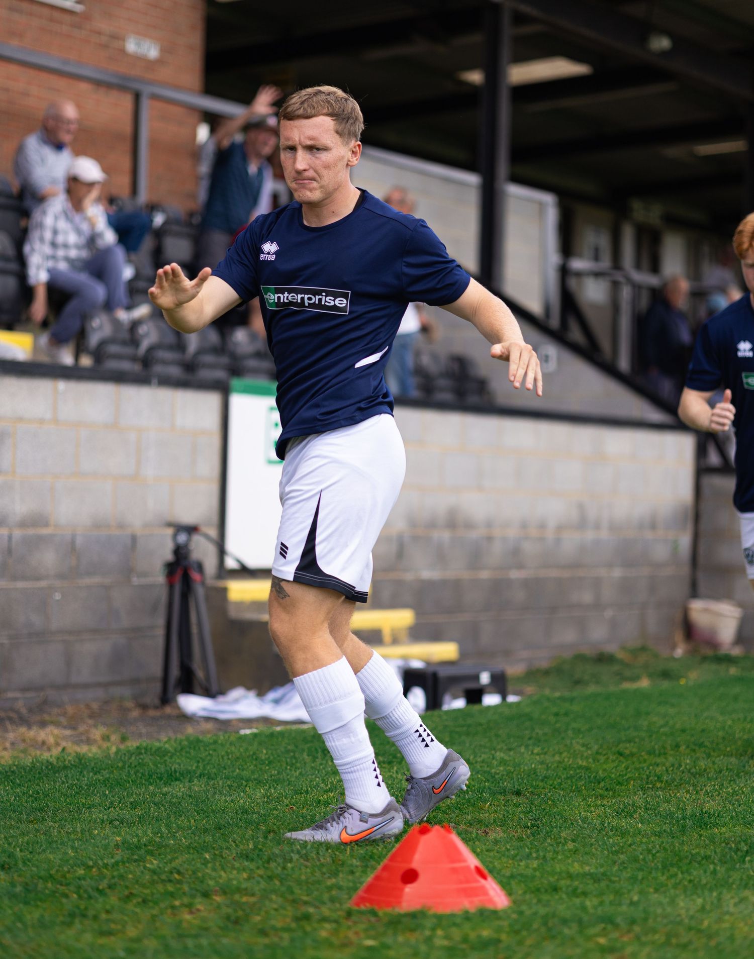A male soccer player in navy blue kit dribbles around orange cones during training at an outdoor stadium.