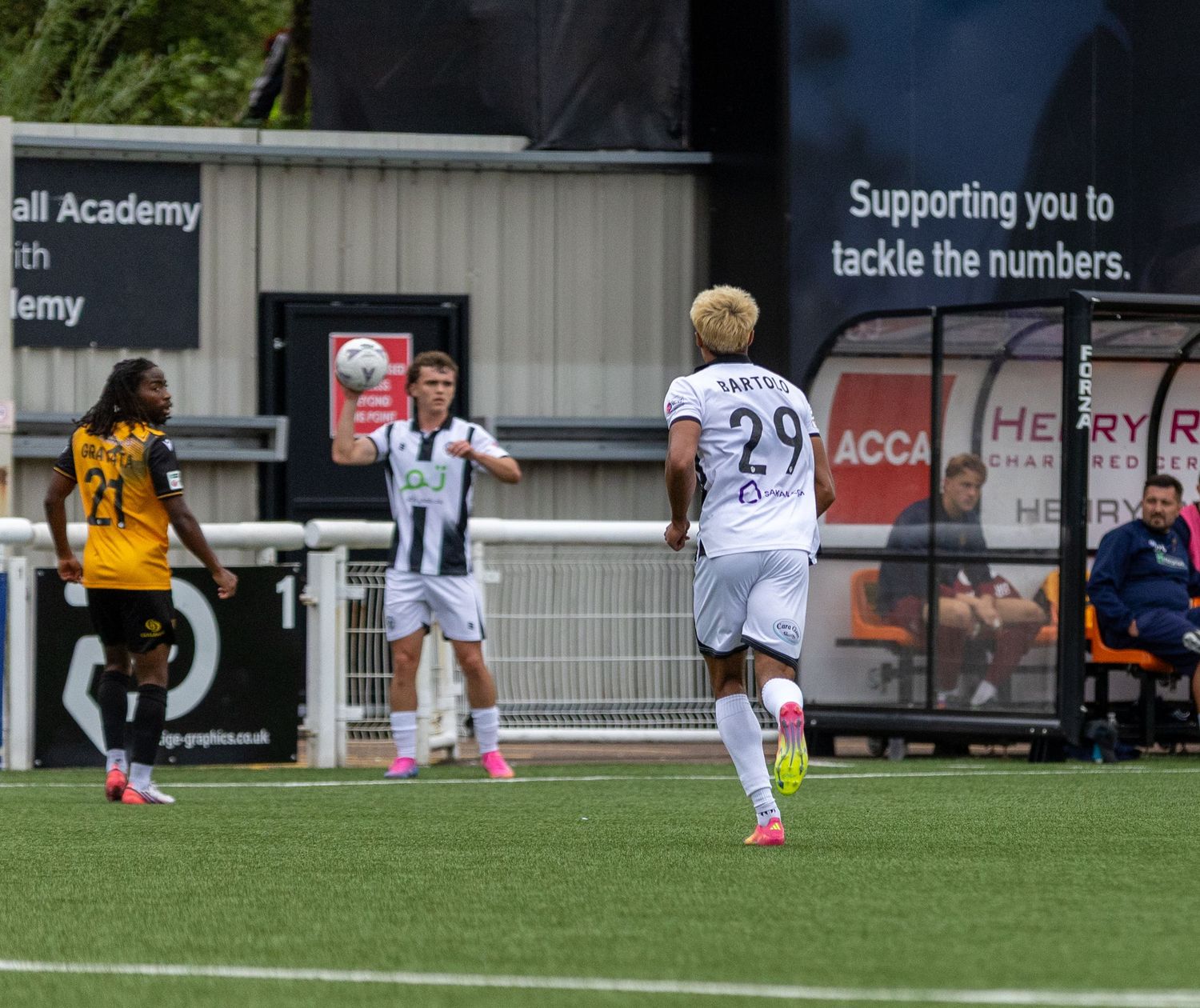 Football players in black and white kits compete on a pitch, with number 29 visible, near an advertising hoarding.