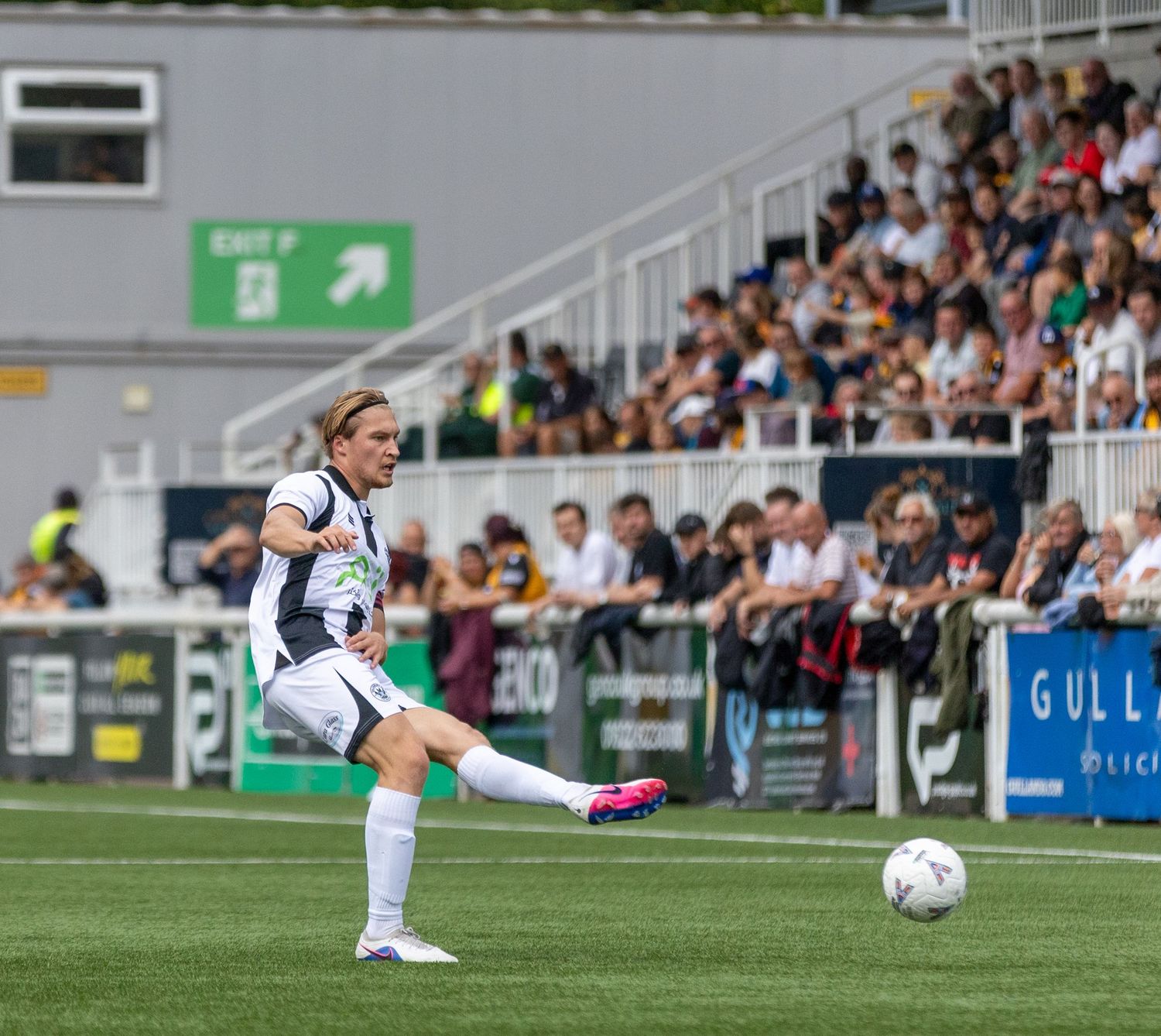 Female soccer player in white kit kicking ball on green pitch with crowded stadium stands in background.
