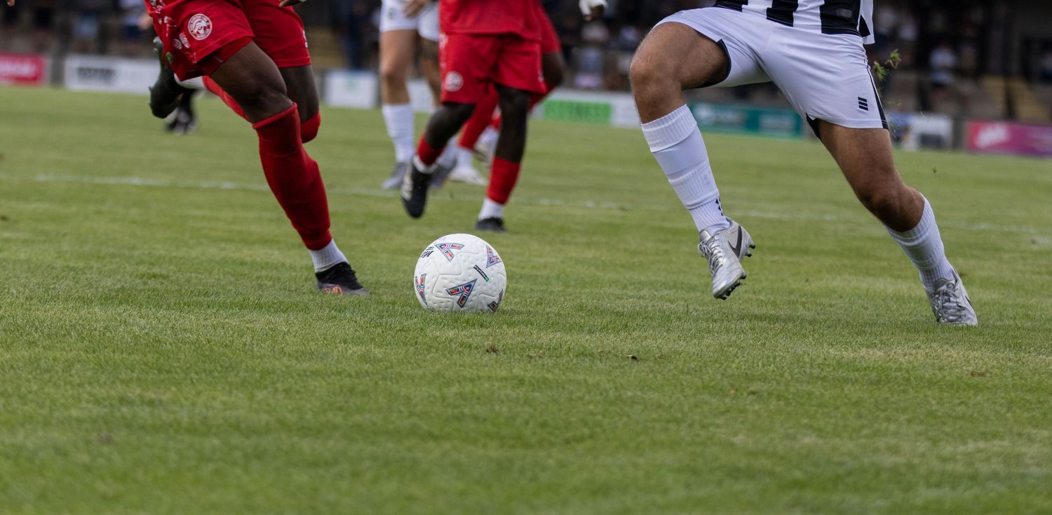 Players in red and black-and-white kits compete for the ball on a green football pitch.