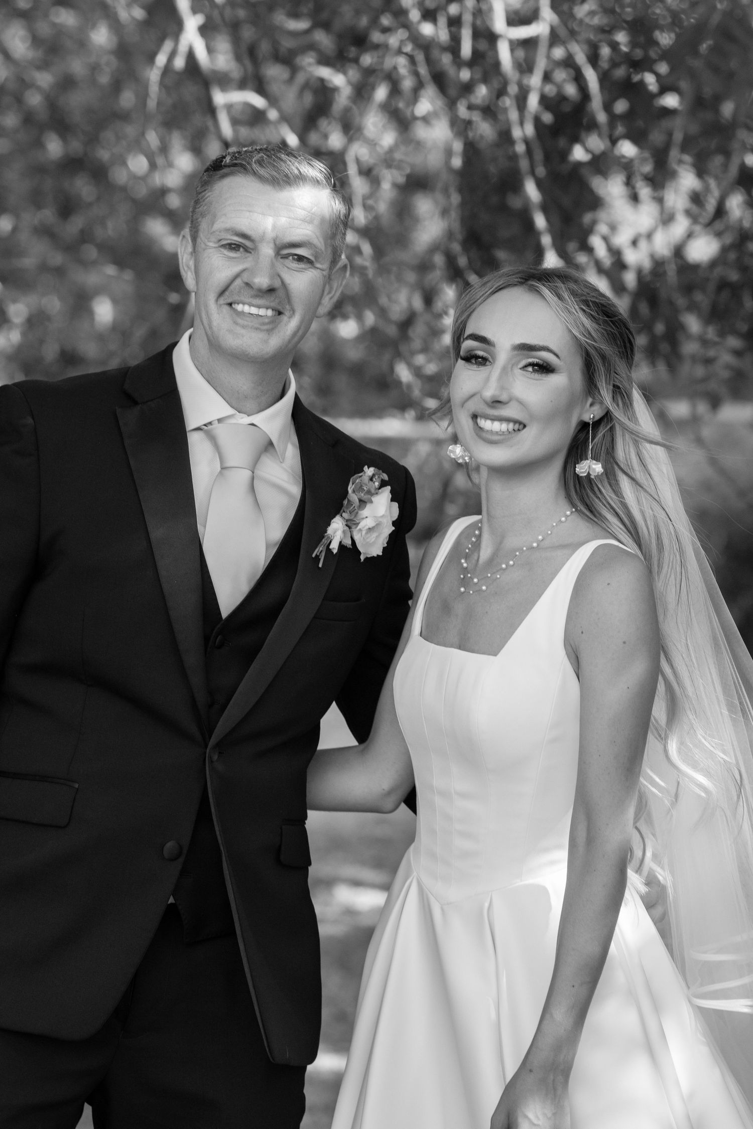 Black and white photo of smiling groom in suit with boutonniere beside bride in white wedding dress.