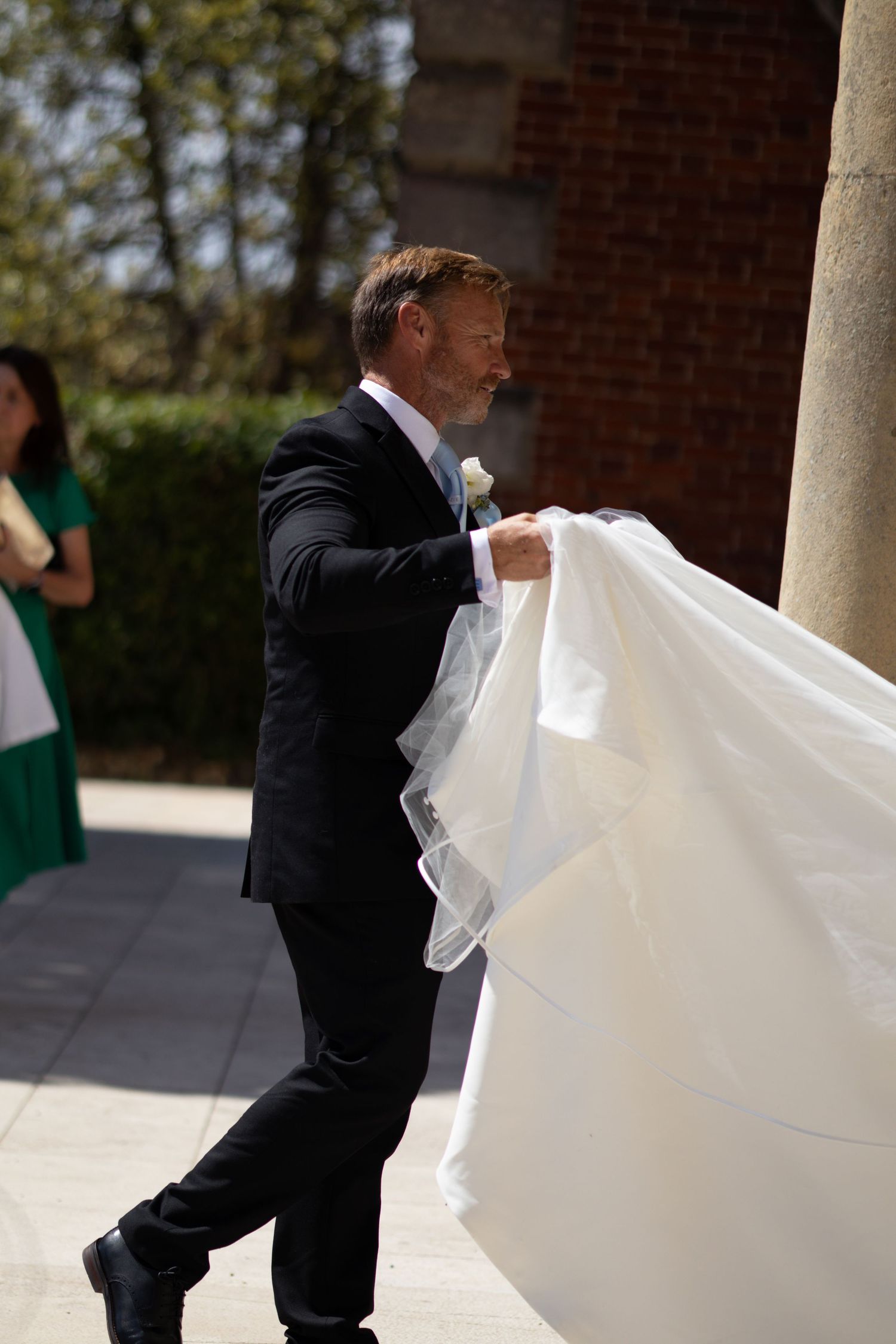 Groom in dark suit lifting and arranging white bridal gown train outside church on wedding day.