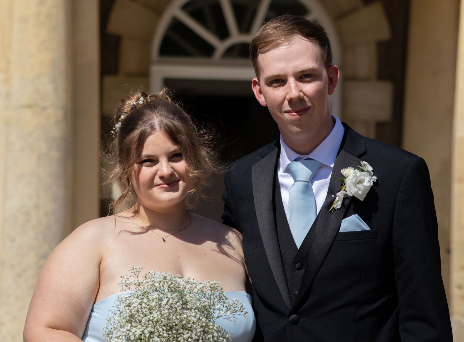 Smiling couple poses outside a grand building, bride holding a white bouquet with baby's breath.