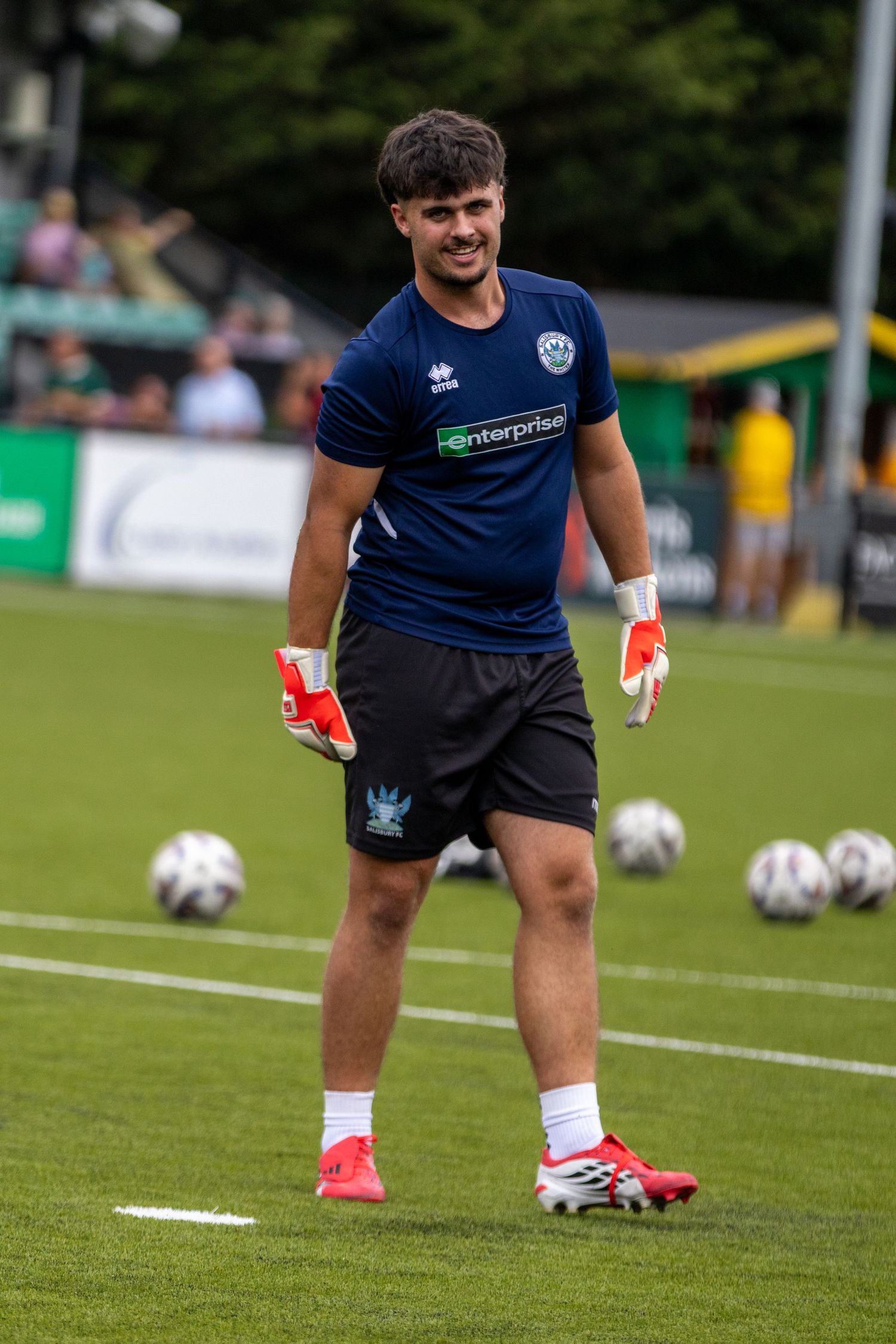 Smiling goalkeeper in navy Enterprise-sponsored kit holds gloves during pre-match warmup on green football pitch.
