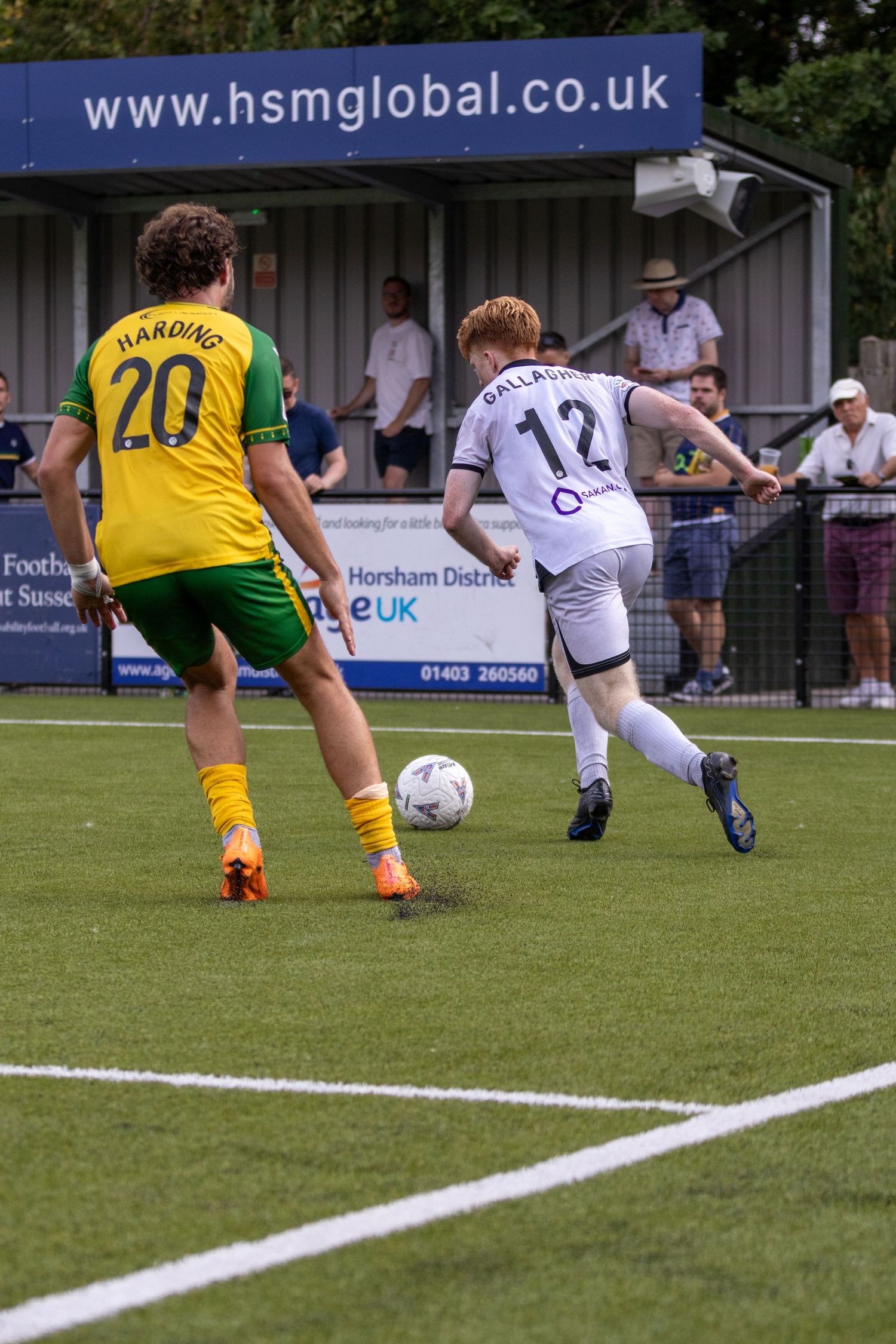 Gallagher number 12 in white shoots past Harding number 20 in yellow during an outdoor football match.