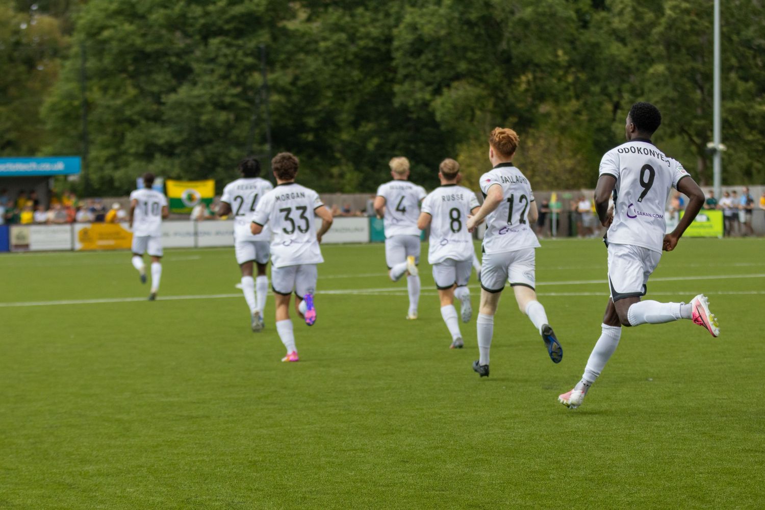 Football players in white kits sprint across a lush green pitch during a match.