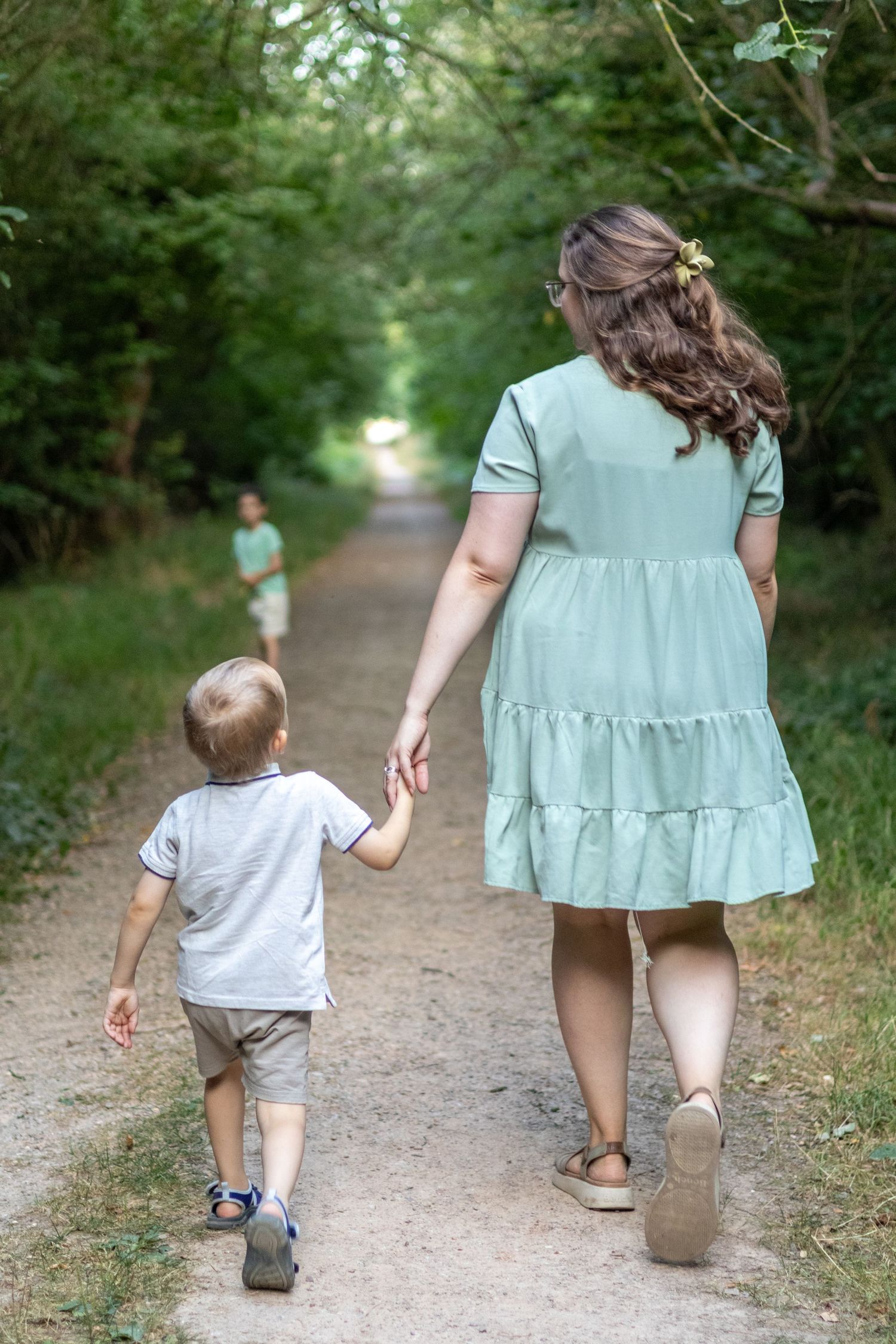 A mother in a mint tiered dress walks with her toddler on a lush green forest trail.