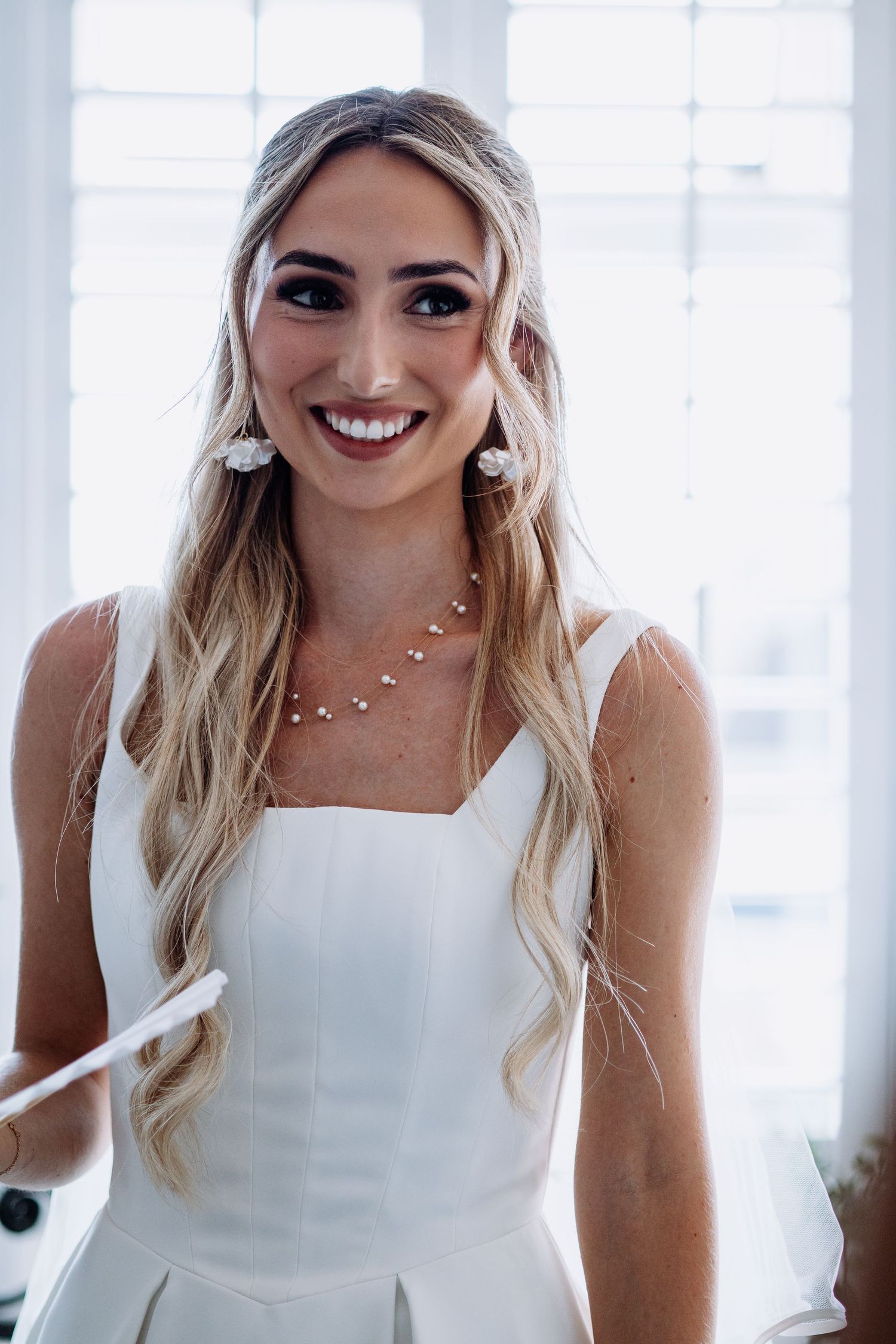 Radiant bride in white dress with long blonde hair wearing pearl earrings and necklace smiling.