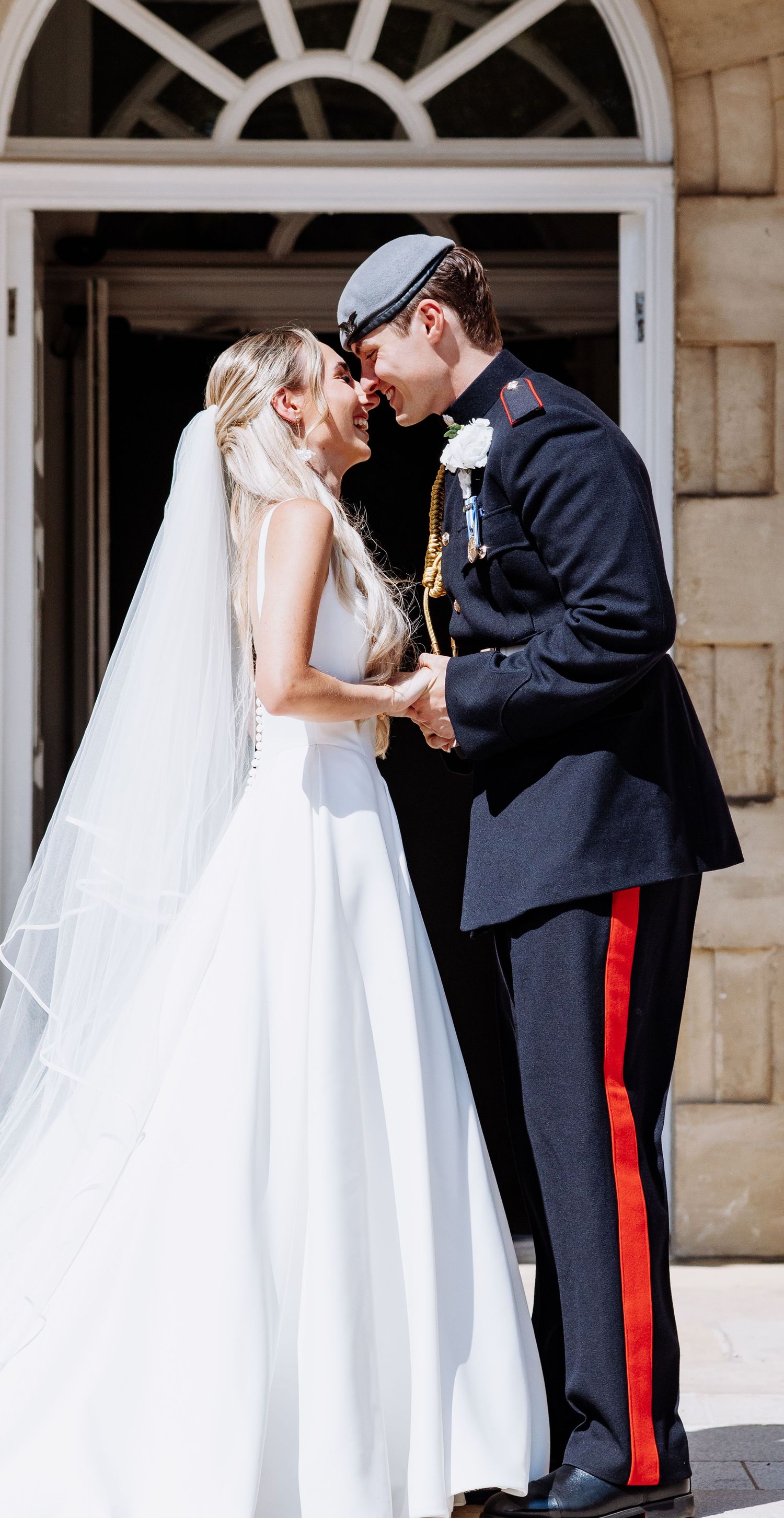 Bride and groom in military uniform touch foreheads romantically outside a grand stone building.