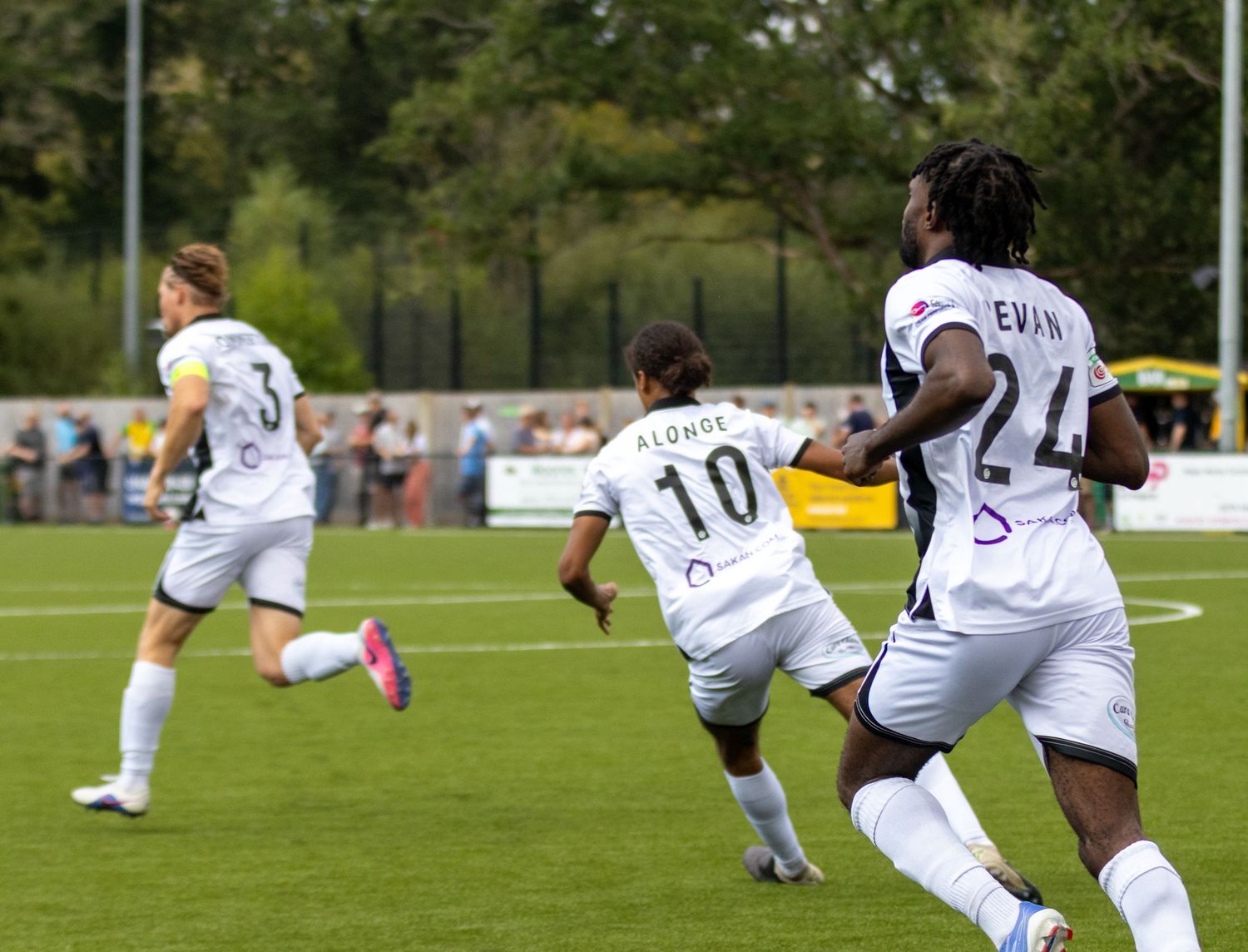 Football players in white kits celebrate a goal on a green pitch during a non-league soccer match.