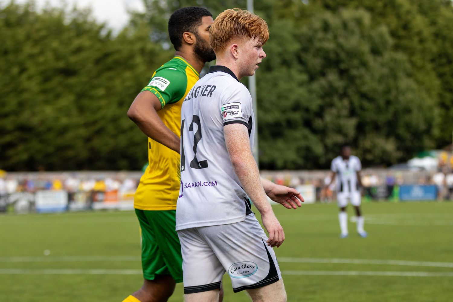 Two footballers, one in yellow-green and one in white number 2 kit, walk side by side on pitch.
