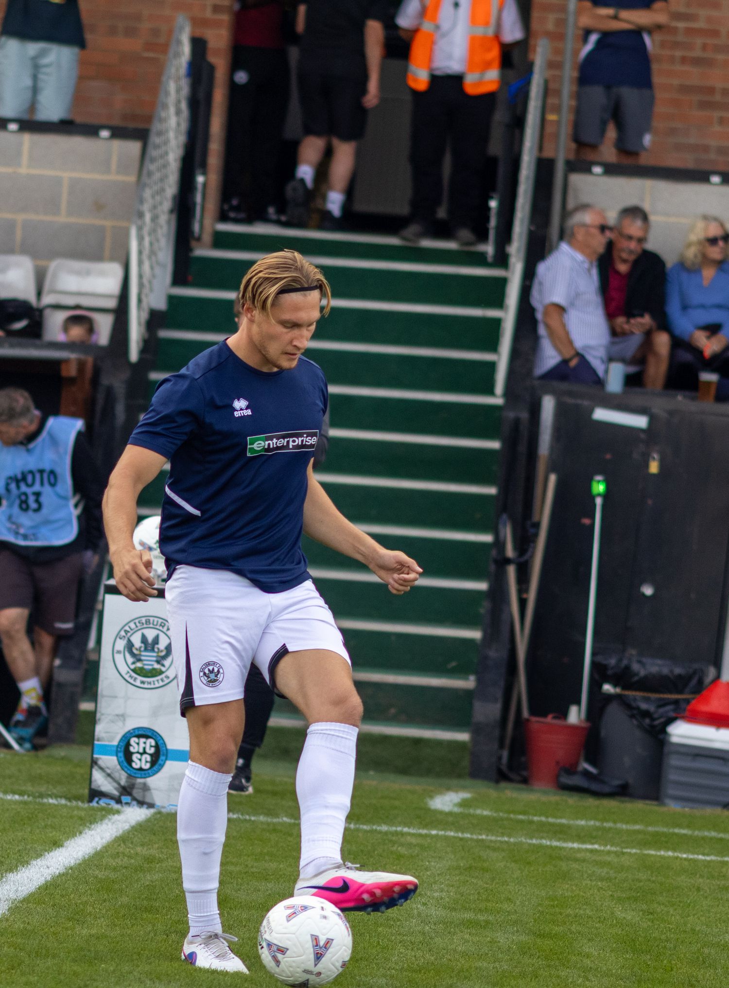 A blond footballer in navy Errea kit controls the ball near the touchline during a football match.