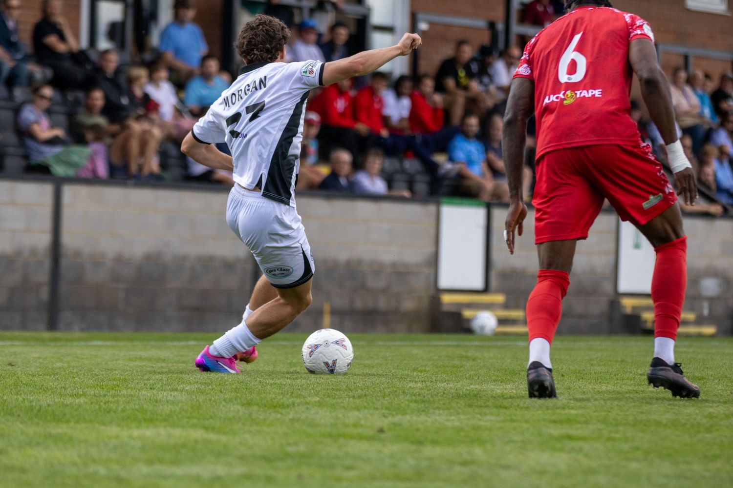 A white-kitted footballer named Holigan dribbles the ball past a red-kitted opponent during a match.