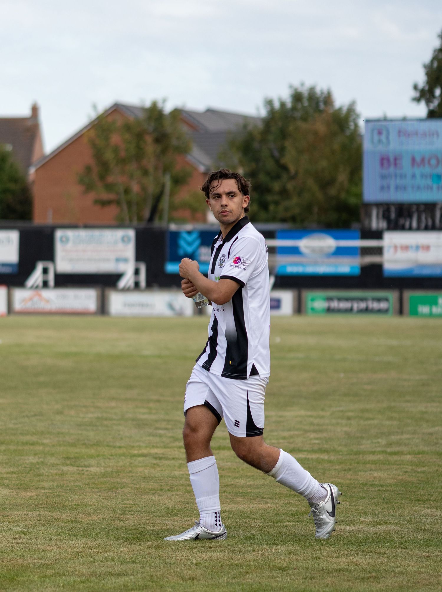 Male footballer in black and white striped kit strides across green pitch during a football match.