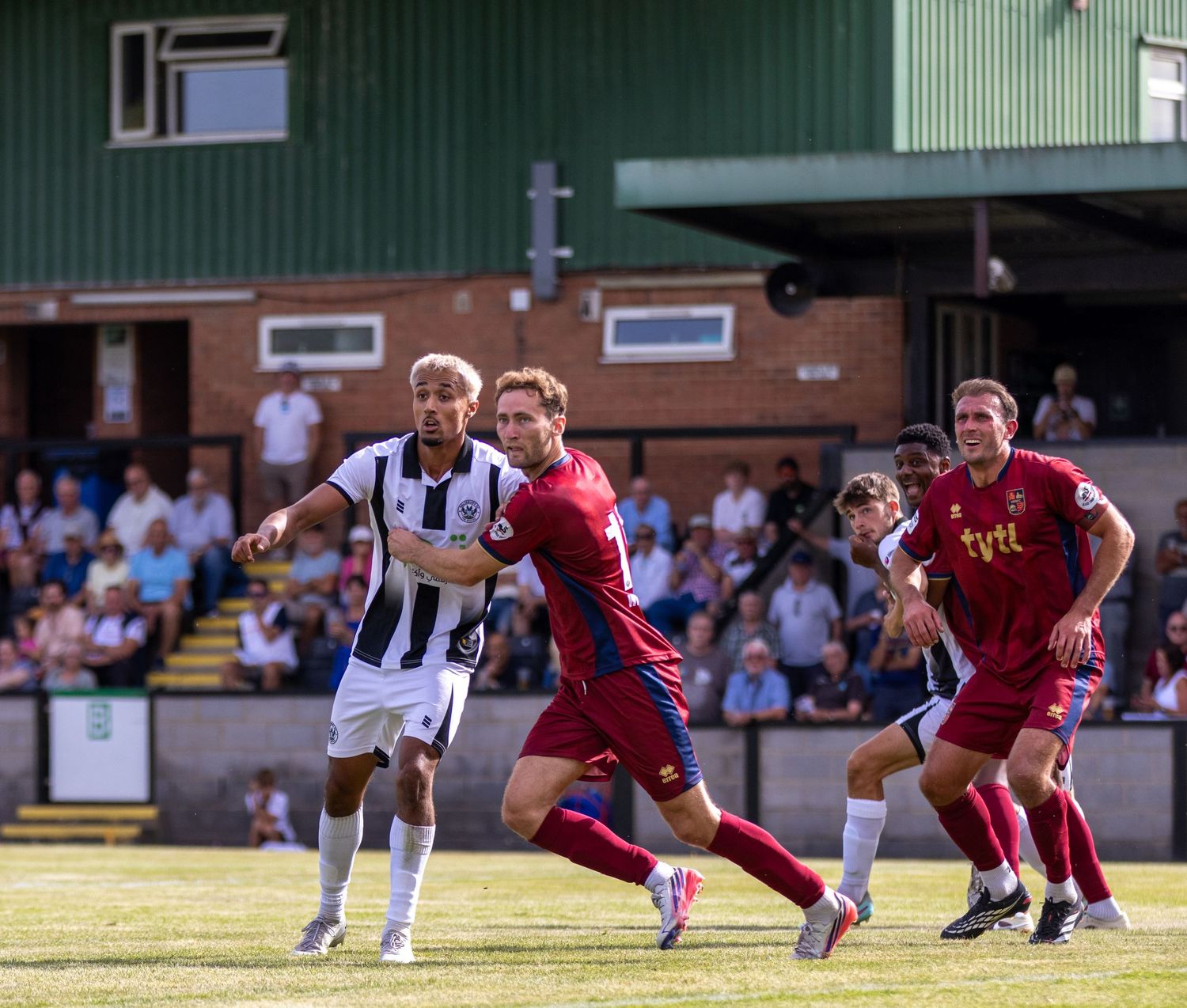 Players in black-white and red kits battle for the ball during a non-league football match in sunshine.
