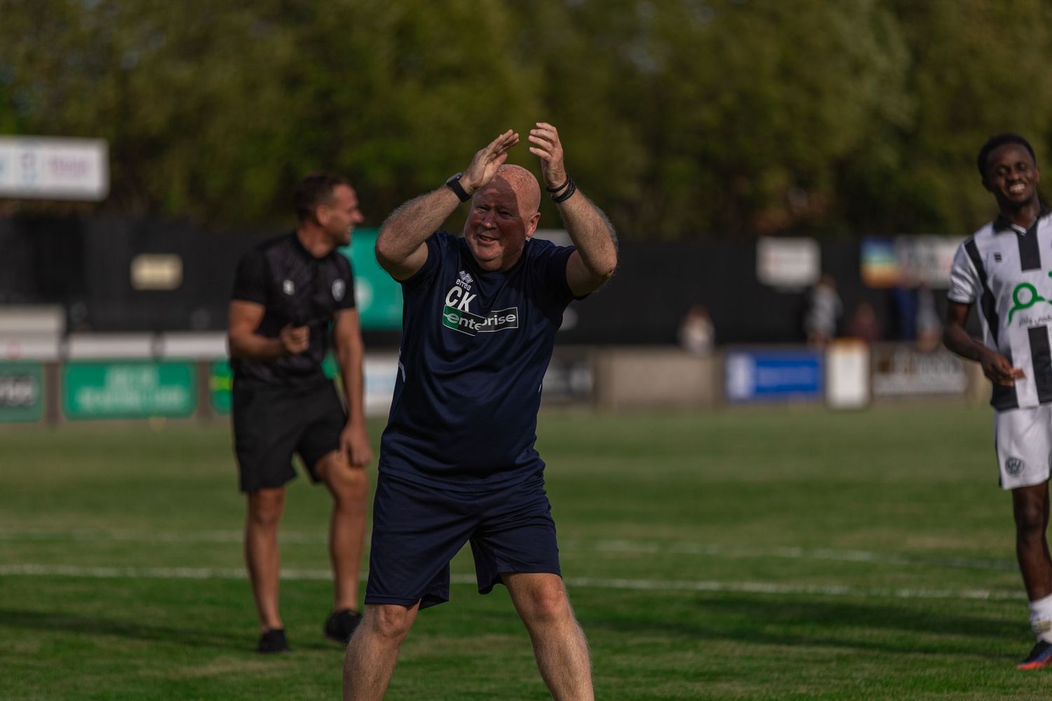 A bald man in a navy shirt reacts emotionally on a football sideline, with players and green trees in the background.