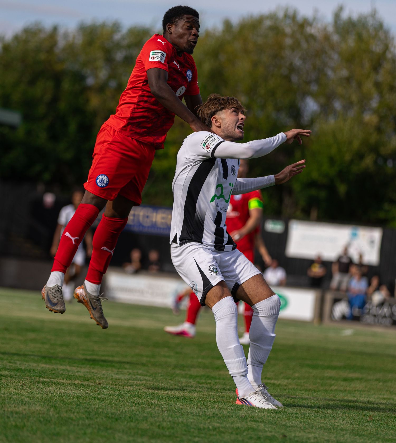 Two soccer players compete for a header during a sunny outdoor match, one in red and one in black and white striped kit.