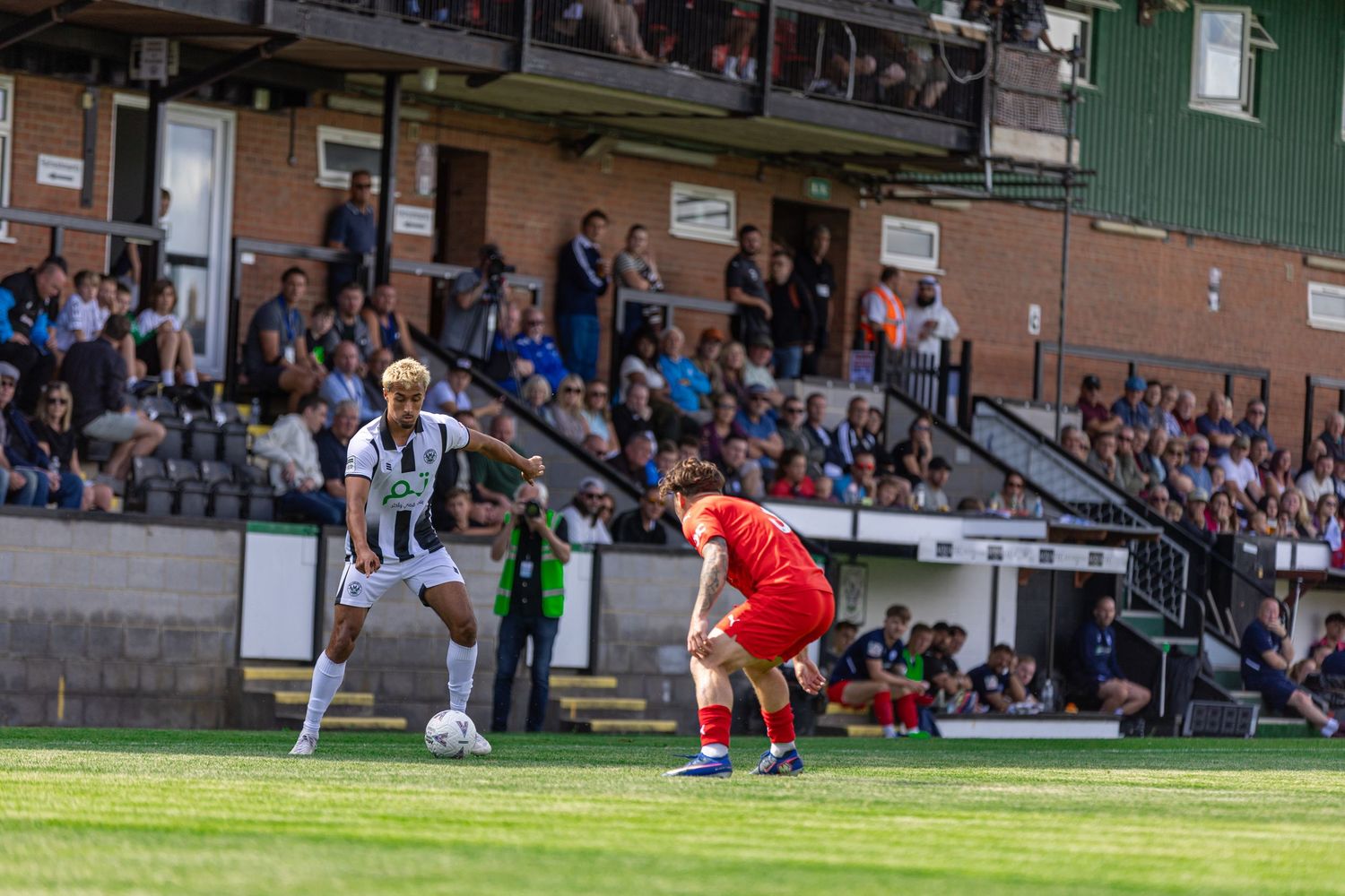 Two soccer players compete on a green pitch with a crowded stadium stand in the background during a sunny match.