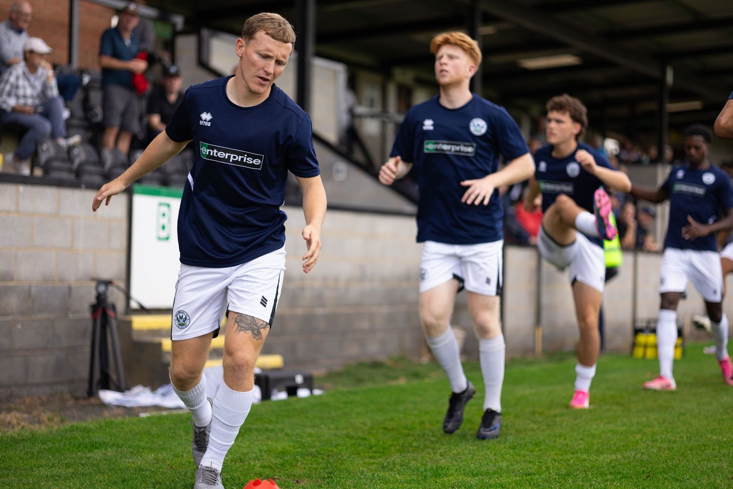 Soccer players in navy blue uniforms warm up on a green pitch before a match at an outdoor stadium.