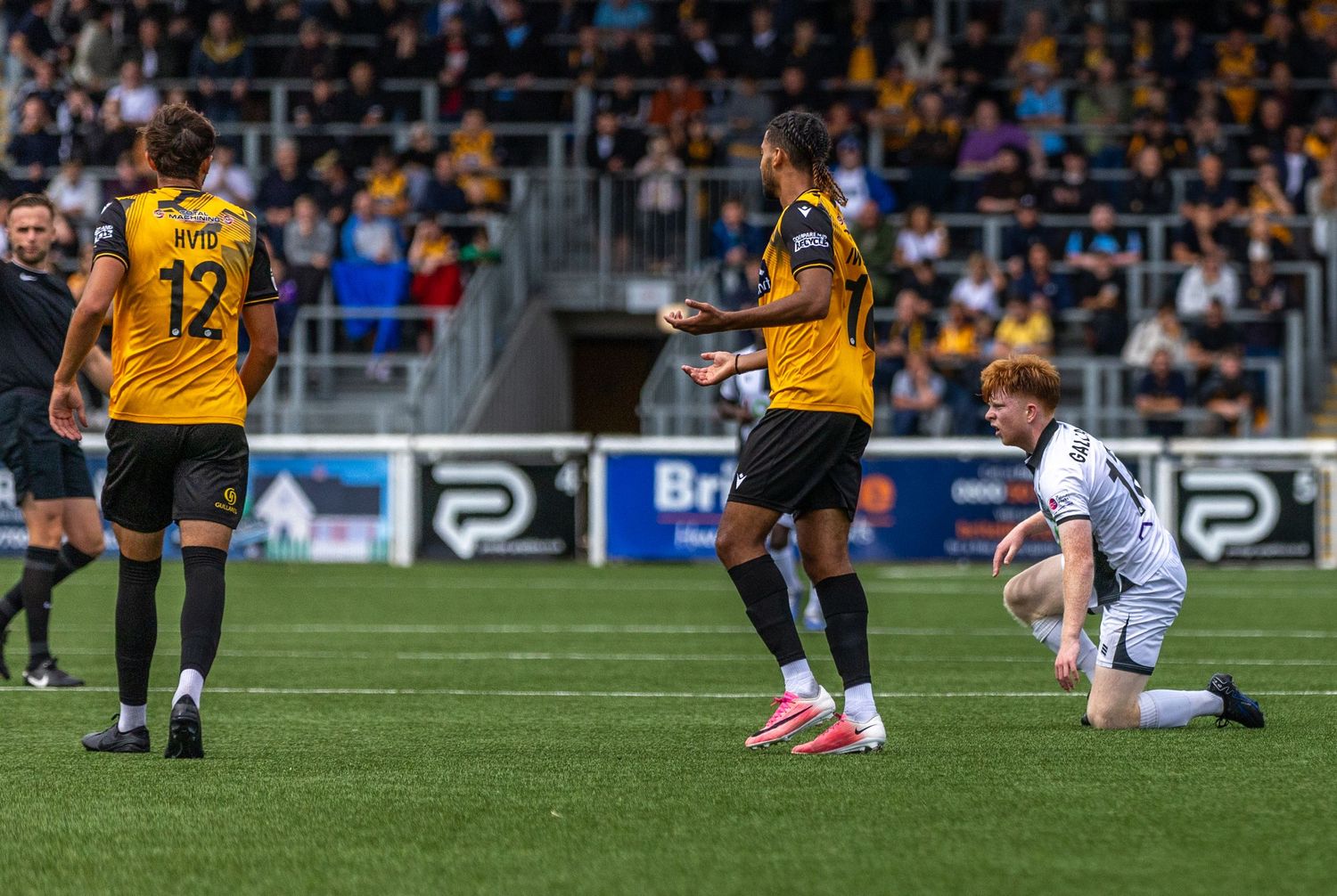Soccer players in gold and black jerseys compete on a stadium pitch, with one player kneeling on the ground.