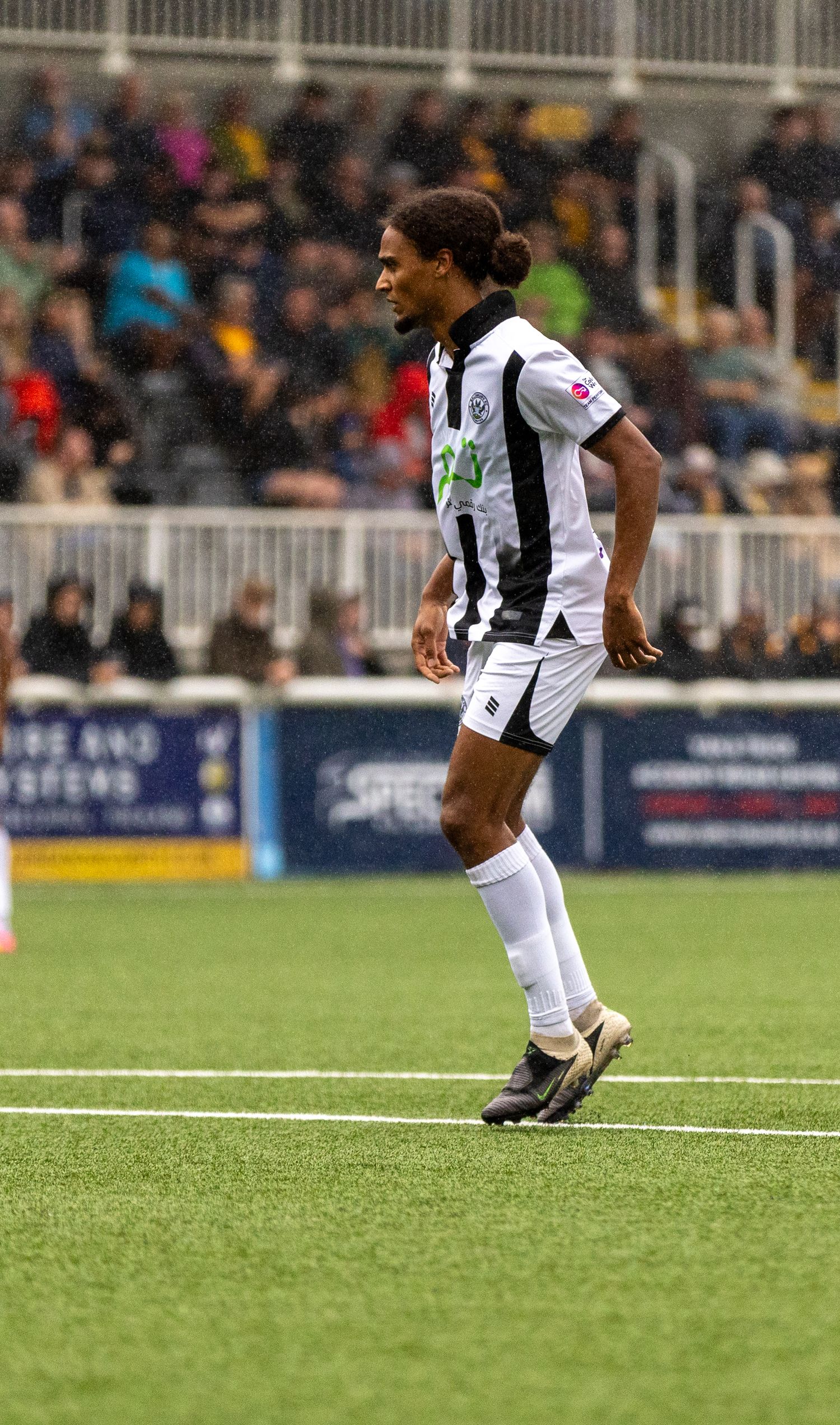 Soccer player in black and white kit walking on green pitch during a match with spectators in background.